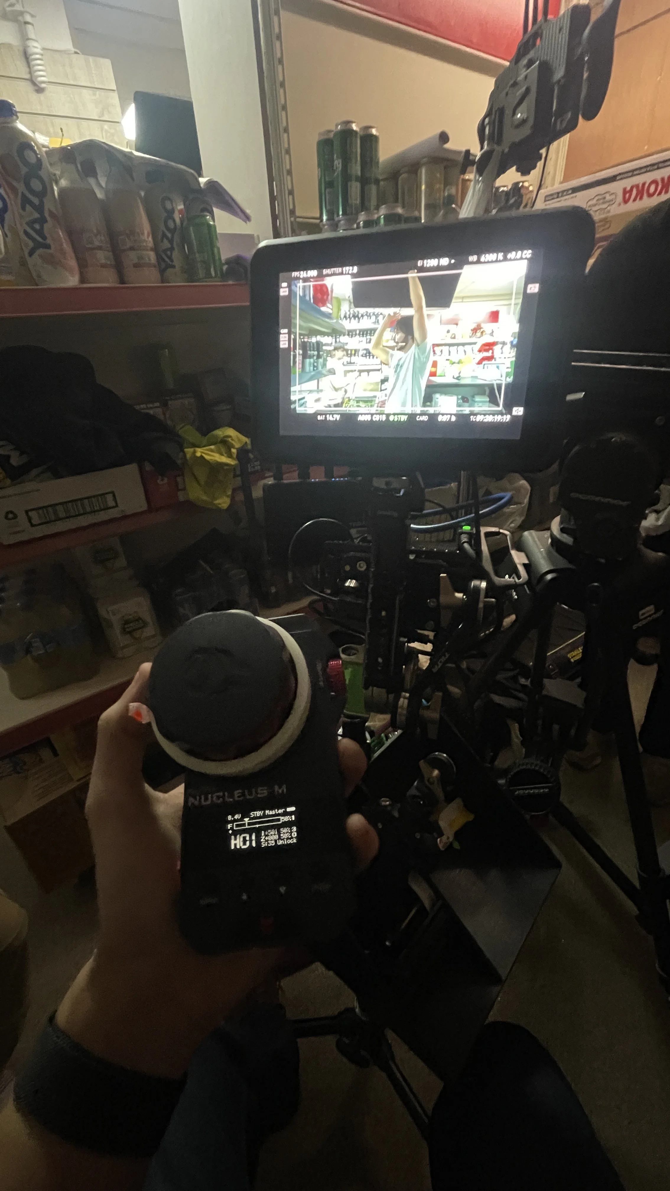 A person operating a film camera on a tripod in a store aisle with shelves of cleaning products and canned goods.