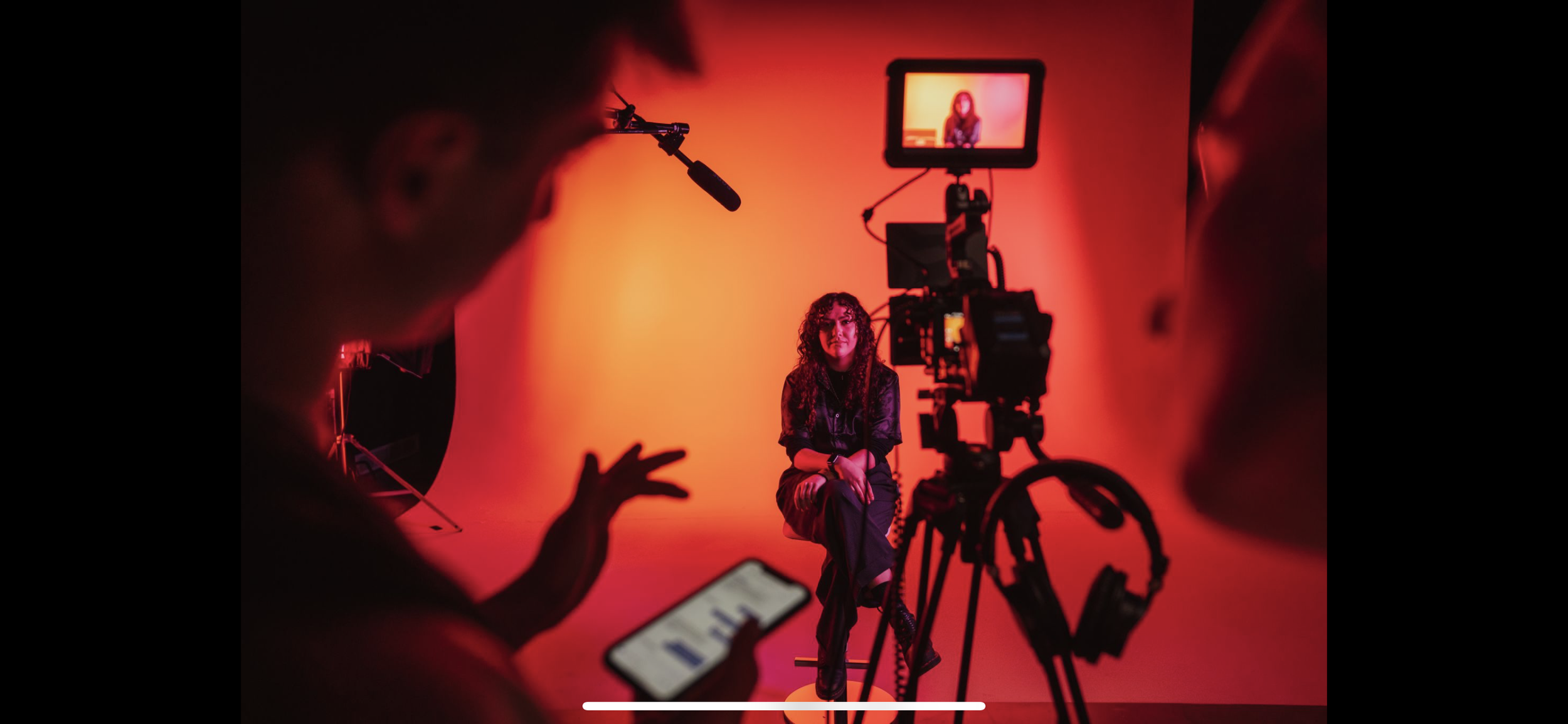 A woman is sitting on a stool in front of a red backdrop during a video recording session. A camera captures her image, which is displayed on a monitor. Crew members are visible in the foreground, with one holding a smartphone and others operating eq