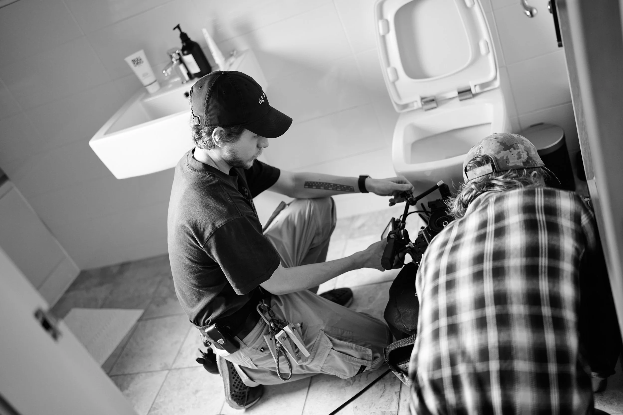 Two men, one wearing a baseball cap and another wearing a camouflage cap, working together on a camera or film equipment in a bathroom with a toilet, sink and toiletries visible.