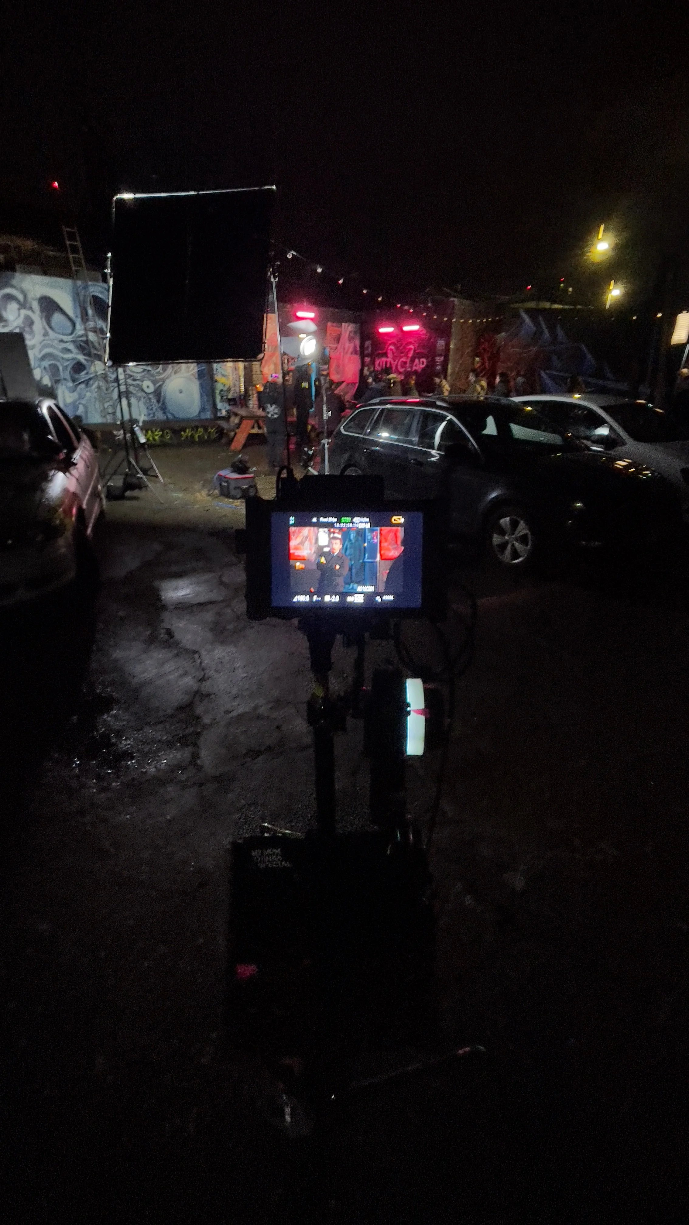 Night scene on a film set showing a camera monitor in the foreground, and equipment, cars, and a graffiti-covered wall in the background, with a person possibly filming or setting up.