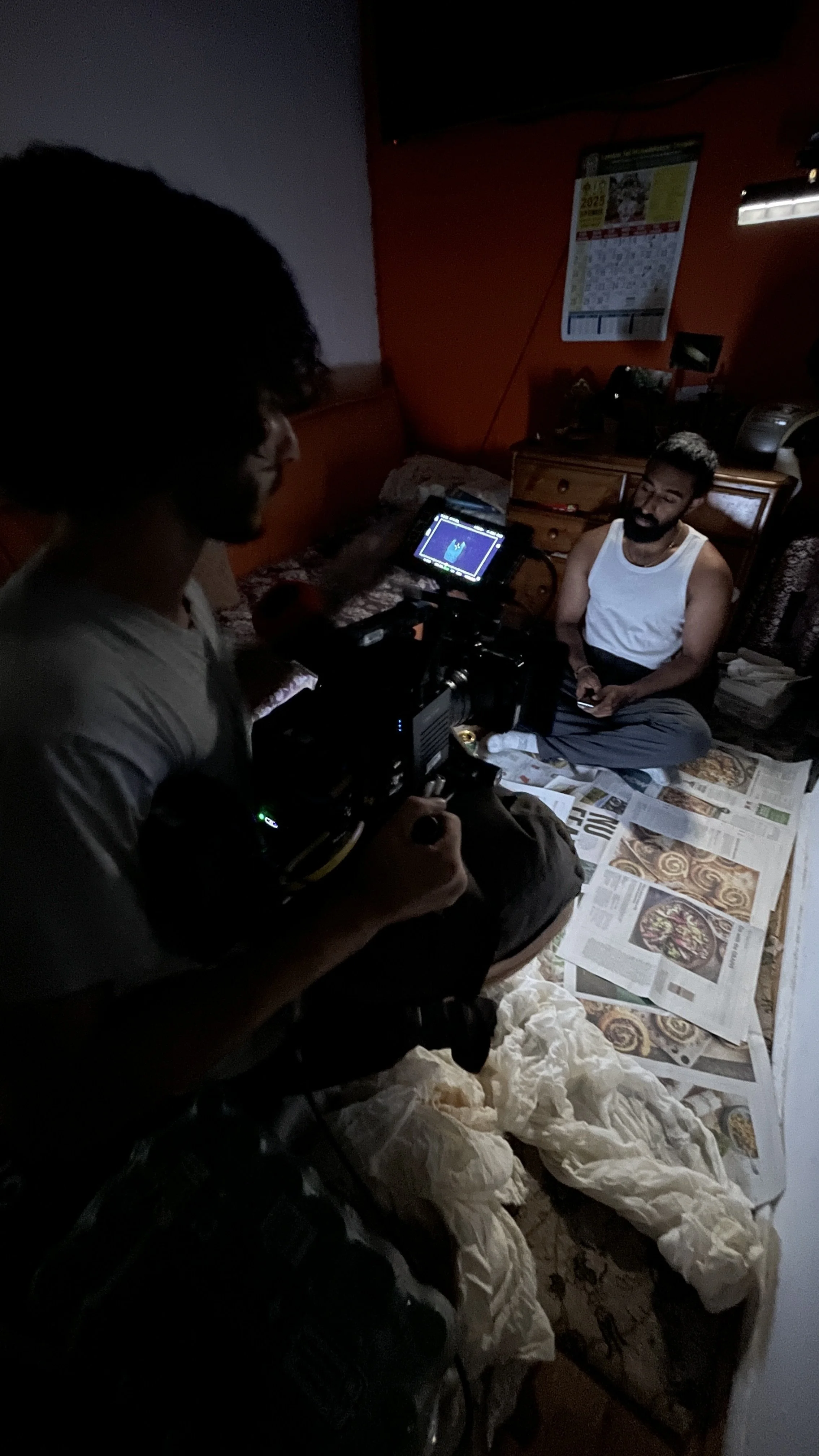 Two men in a dimly lit room sitting on a bed covered with newspapers, with one man holding a camera and the other man sitting cross-legged, smiling, and holding a phone, surrounded by various electronic equipment.