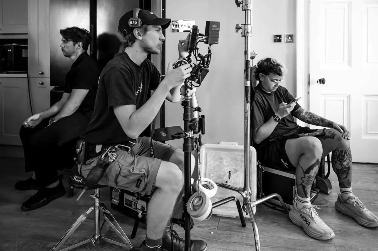 A filming crew on set with one person operating a camera and two people sitting and using their phones.