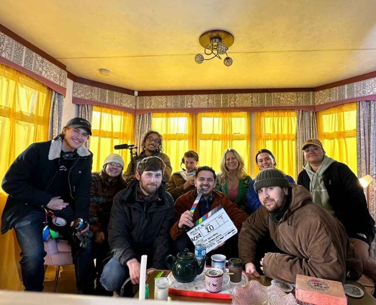 Group of people smiling and posing for a photo in a room with yellow curtains, with one person holding a clapperboard indicating a film or video production scene.