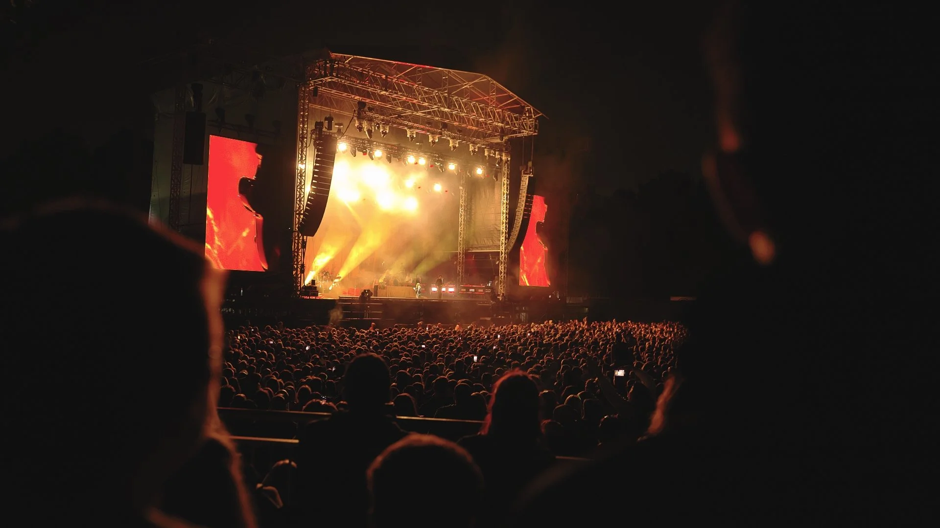 A nighttime outdoor concert stage with bright yellow and orange lights, large screens on either side, and a crowd of people watching. blossoms big bank holiday weekend