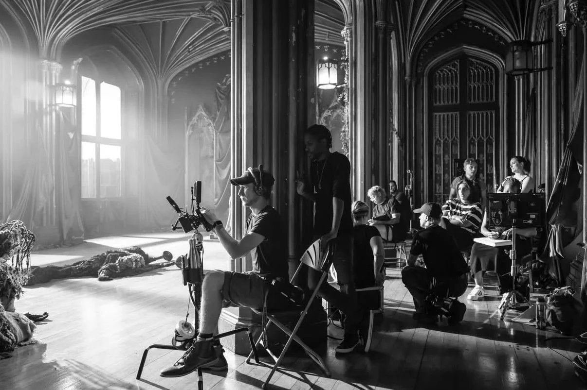 Film crew filming in a grand, ornate room with large windows and tall curtains. Crew members work with equipment, some seated and others standing, preparing for a scene.