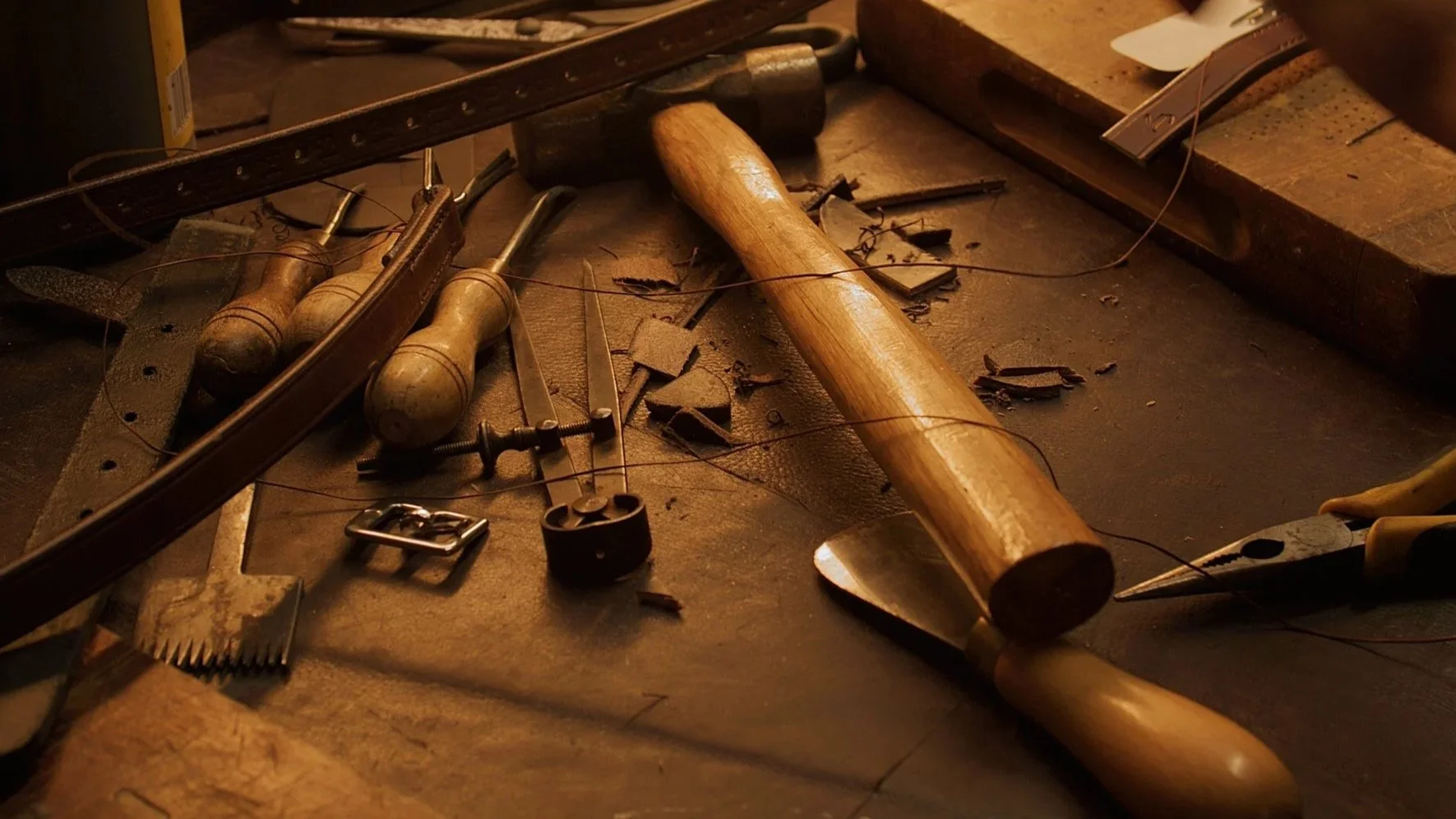 Workstation with woodworking tools including a hammer, screwdrivers, pliers, and metal wire, along with wood shavings and scraps scattered on the surface. Leather saddle making. Saddlery