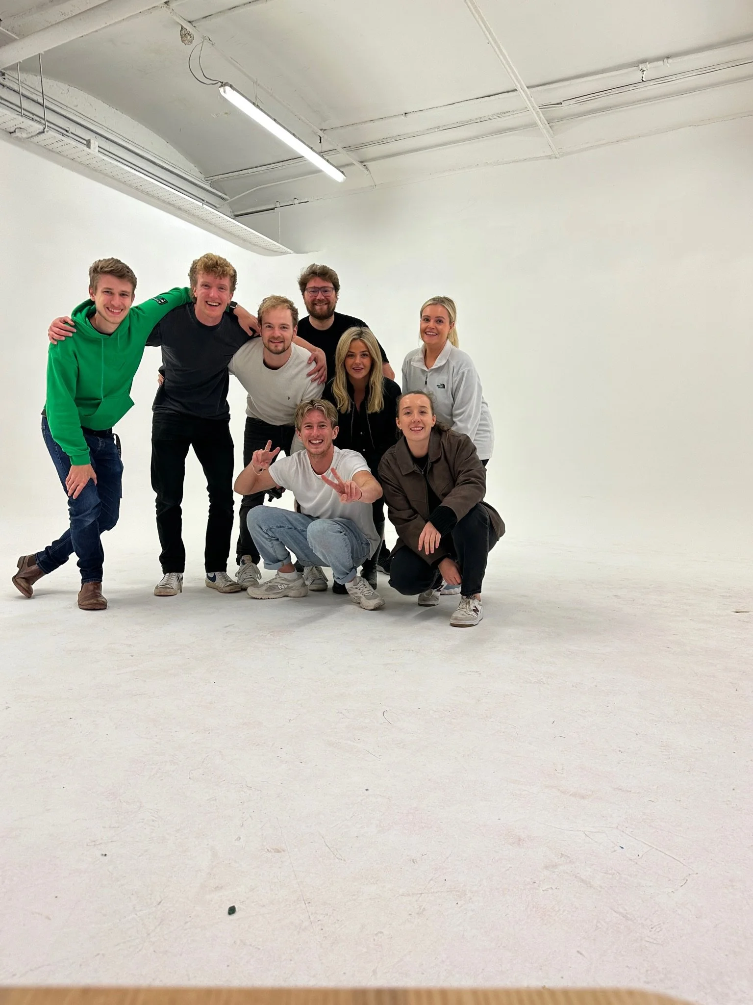 A group of nine young adults posing together in a white studio with minimal background, all smiling and making friendly gestures.