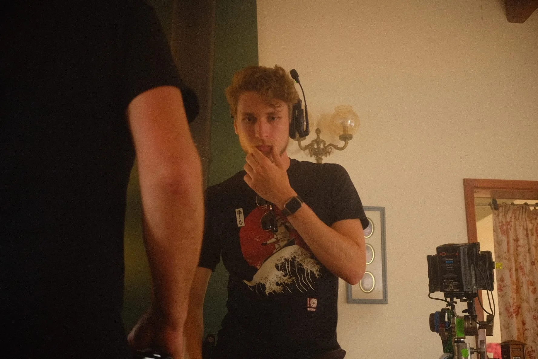 Young man wearing headphones and a black t-shirt with a graphic design, standing in a room with vintage wall sconce, framed mirror, and electronic equipment, looking surprised or questioning.