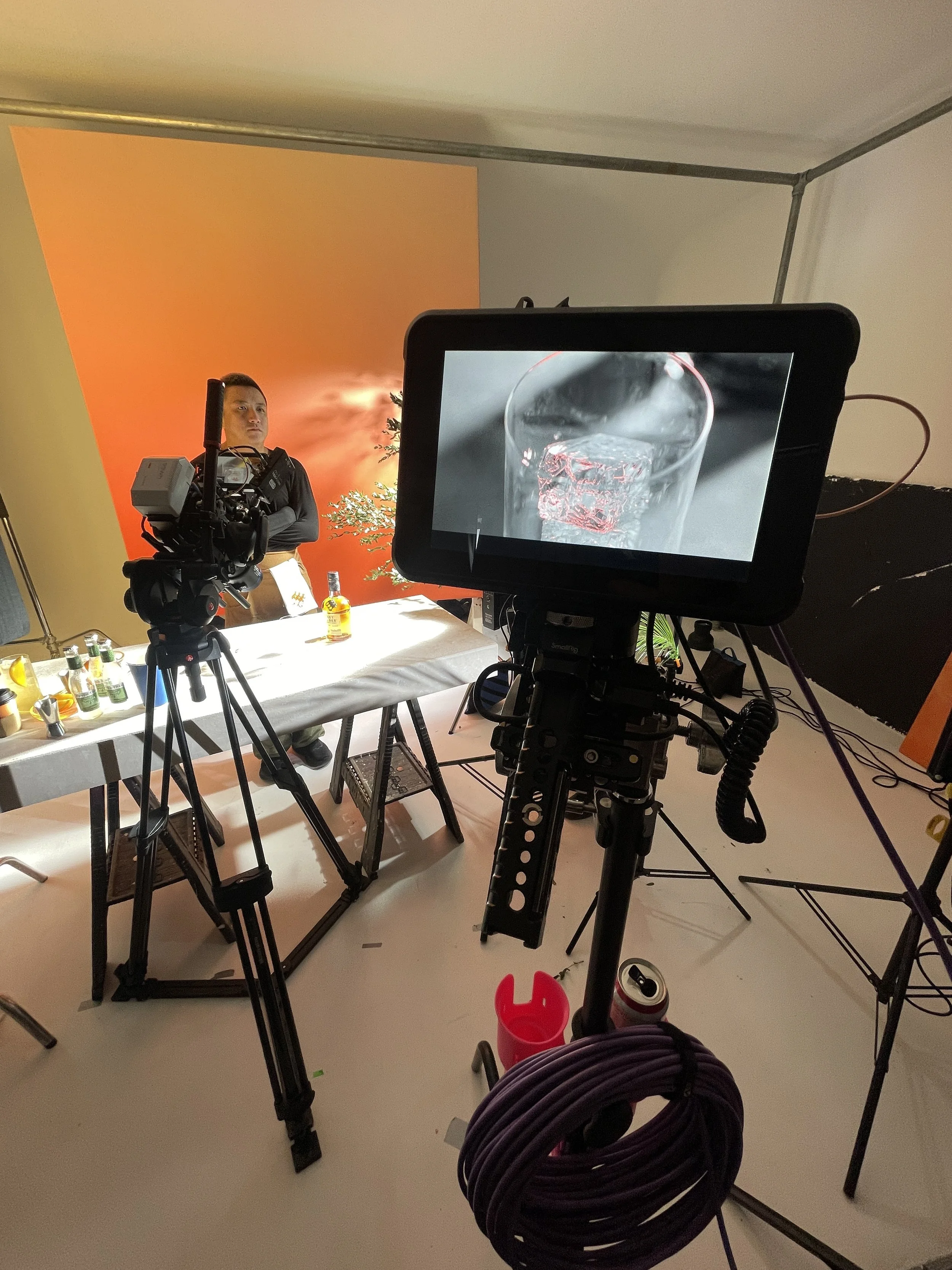 Behind-the-scenes view of a photo or video shoot setup with a person standing in front of an orange backdrop, a camera mounted on a tripod, and an monitor displaying a close-up of a glass with ice water.
