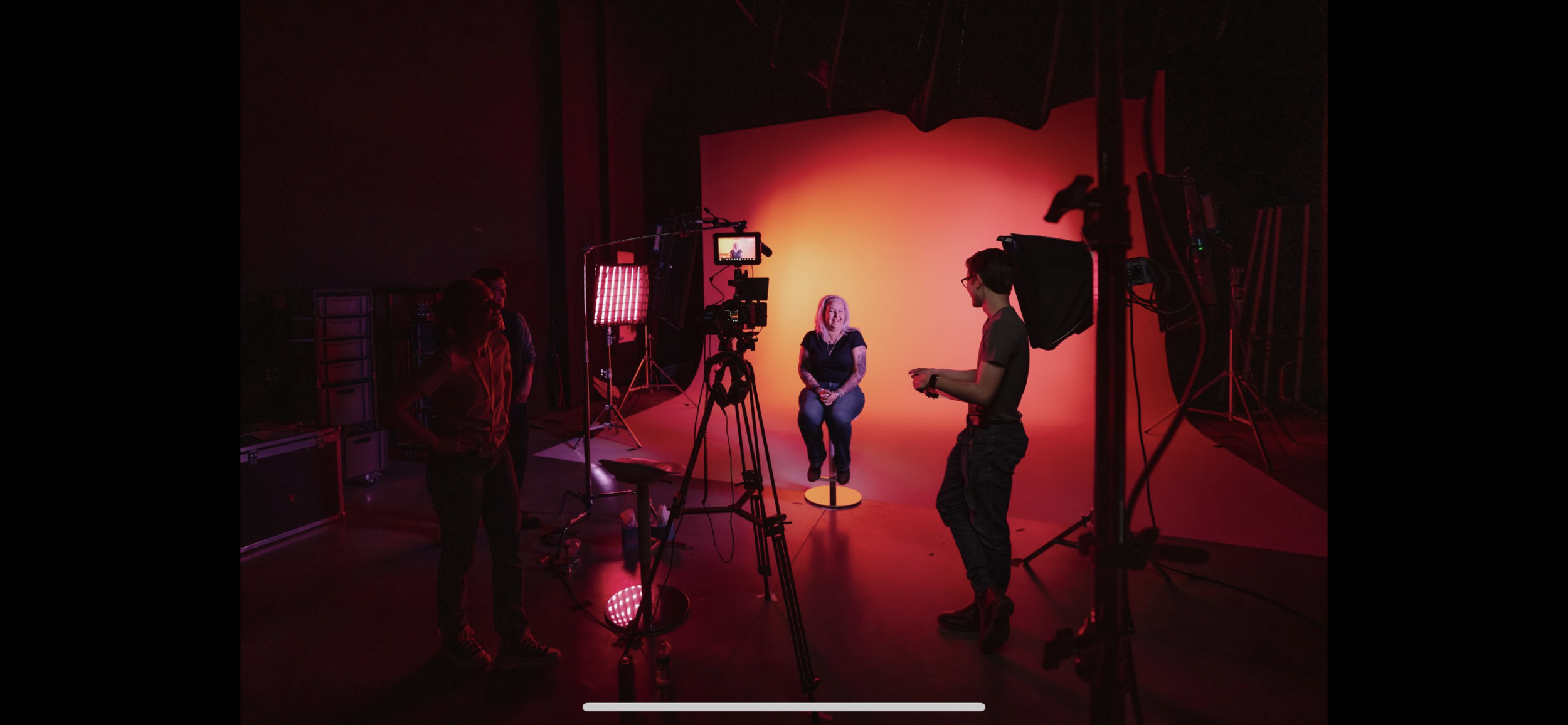 Photographer filming a woman sitting against a warm orange and yellow backdrop in a studio with professional lighting and equipment.