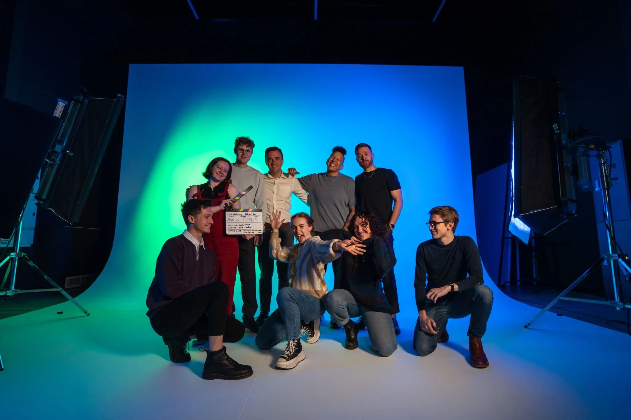 A group of nine young people posing together on a stage, some standing and some sitting, with a colorful blue and green background and studio lighting equipment visible.