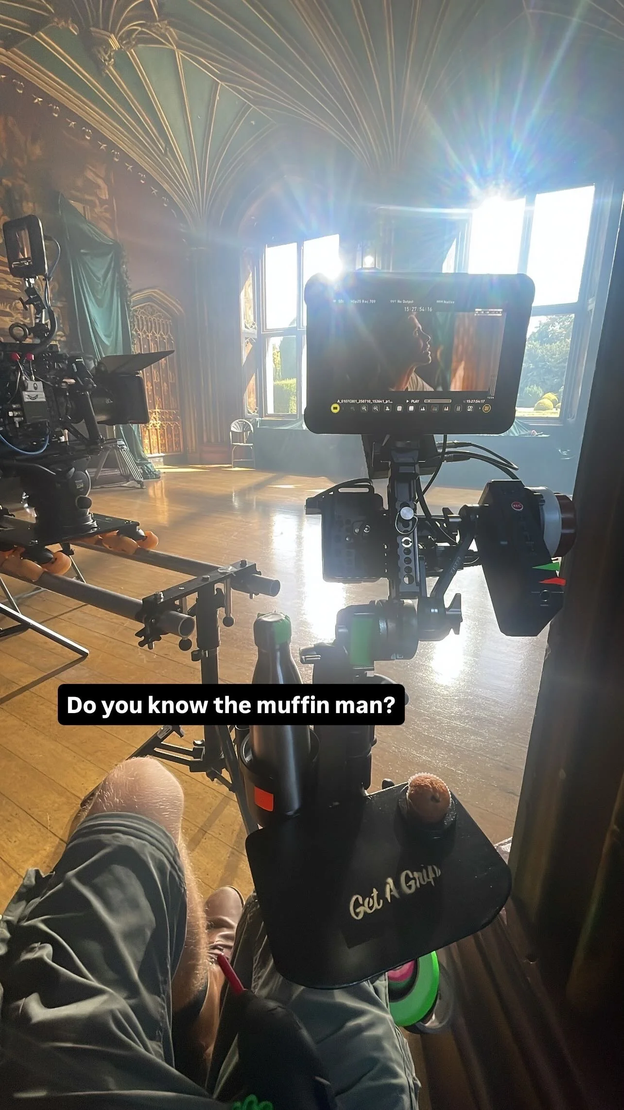 A film set with camera equipment facing a decorated stage with large windows and elaborate ceiling details. The image shows the view from a seated person with crossed legs, holding a muffin, with a caption that says 'Do you know the muffin man?'.