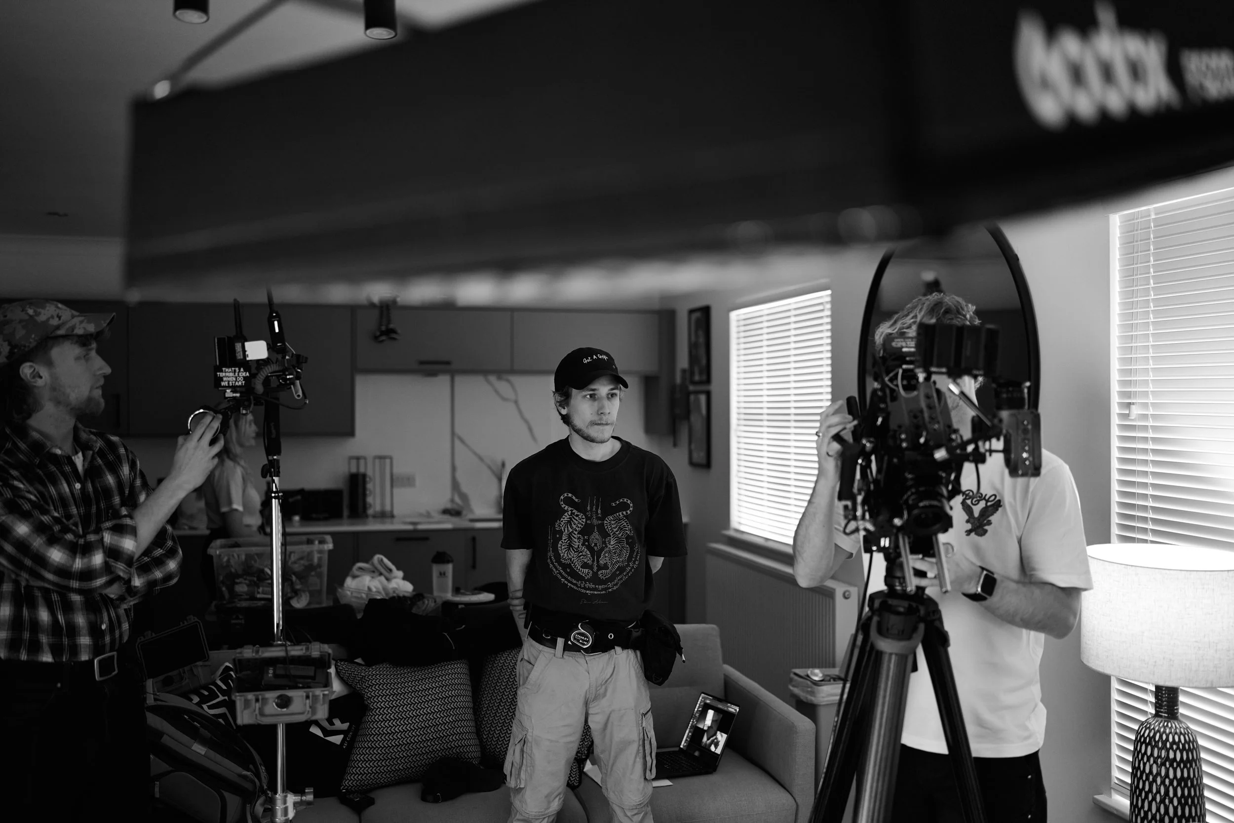 A black-and-white photo of a behind-the-scenes film or photo shoot in a living room. Several crew members are setting up equipment, including a camera on a tripod. One person is adjusting the camera, another is holding a light, and a young man in cas