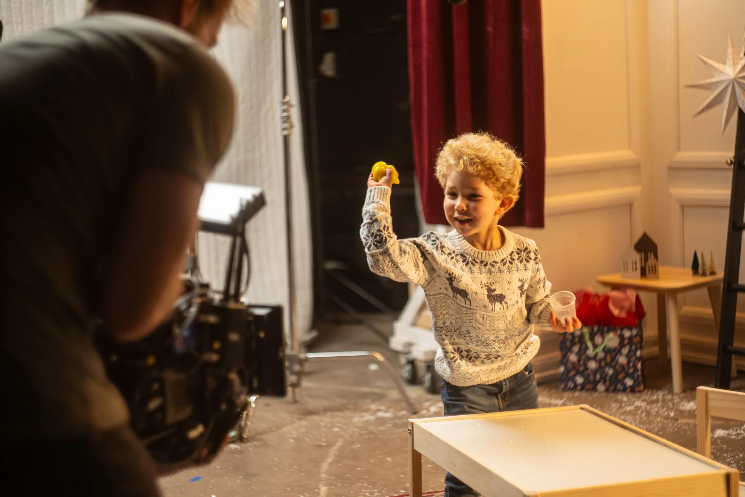 A young boy with curly blond hair wearing a Christmas sweater holding a yellow item and a small container, smiling and interacting with a person who appears to be filming or taking a photo, in a decorated indoor setting.