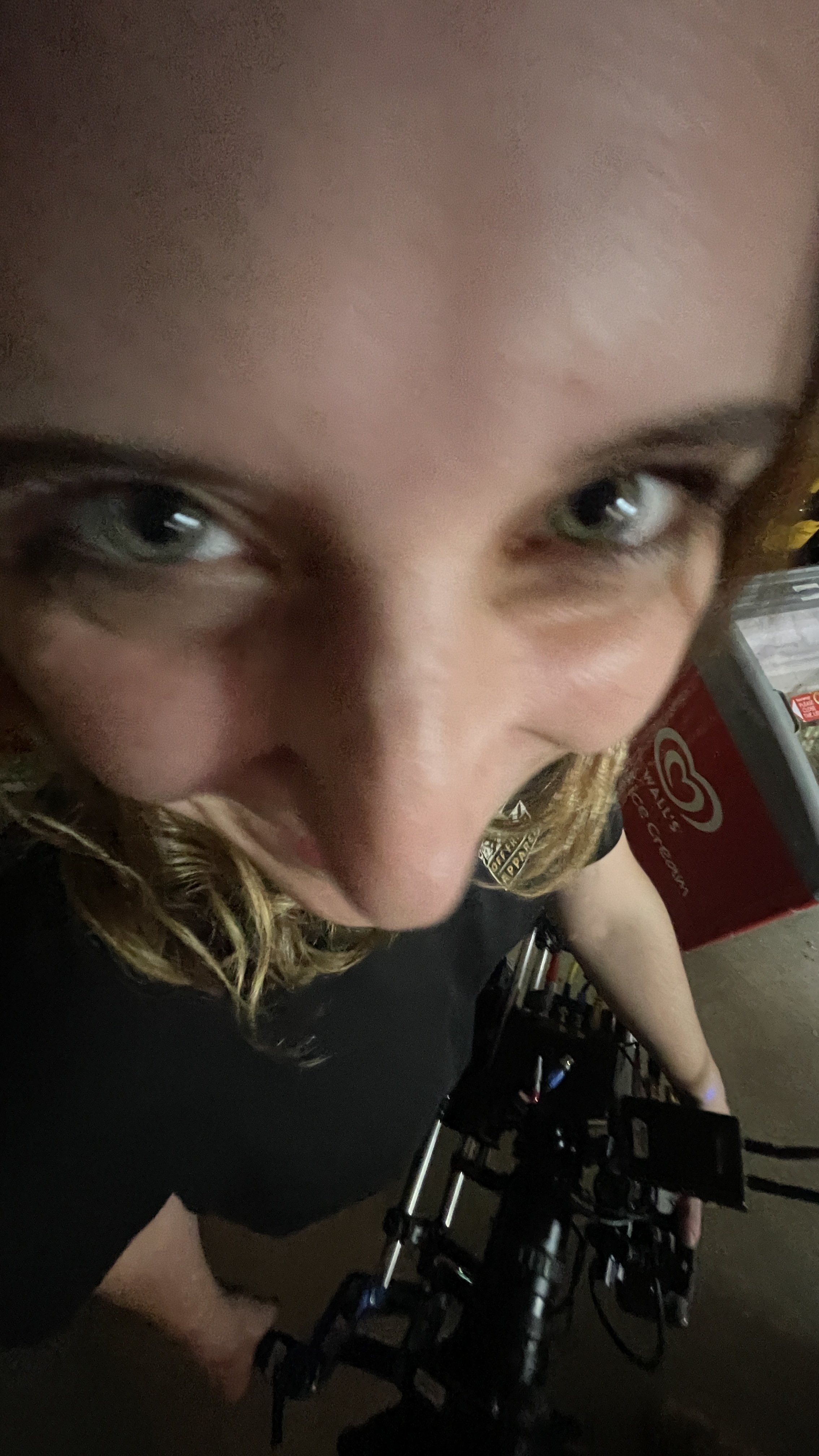 Close-up of a person's face with green eyes and curly hair, holding a camera or phone, with shelves and a red ice cream freezer in the background.