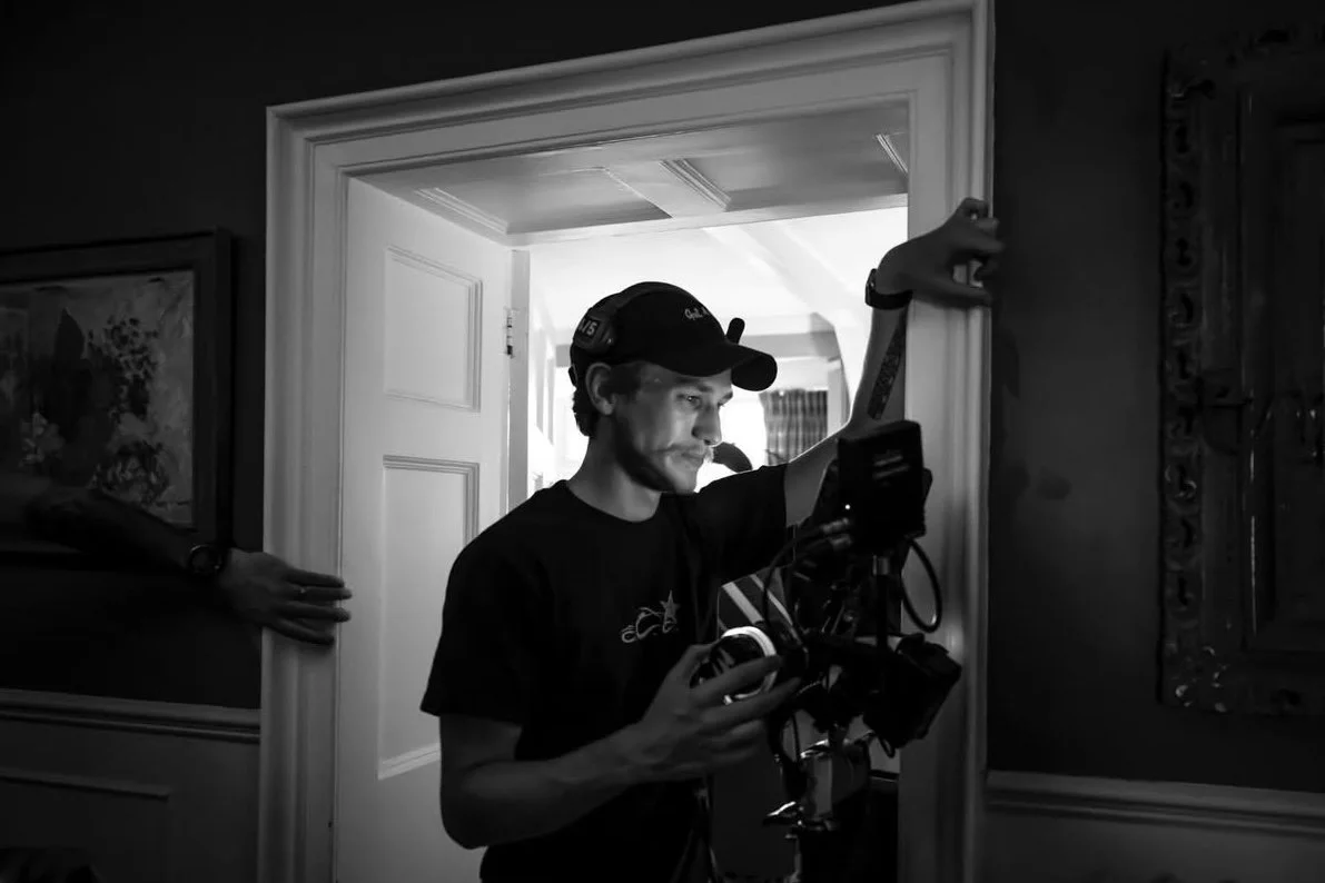 A young man wearing a baseball cap and headphones is operating a film camera indoors, with an open door behind him leading to a lit room.