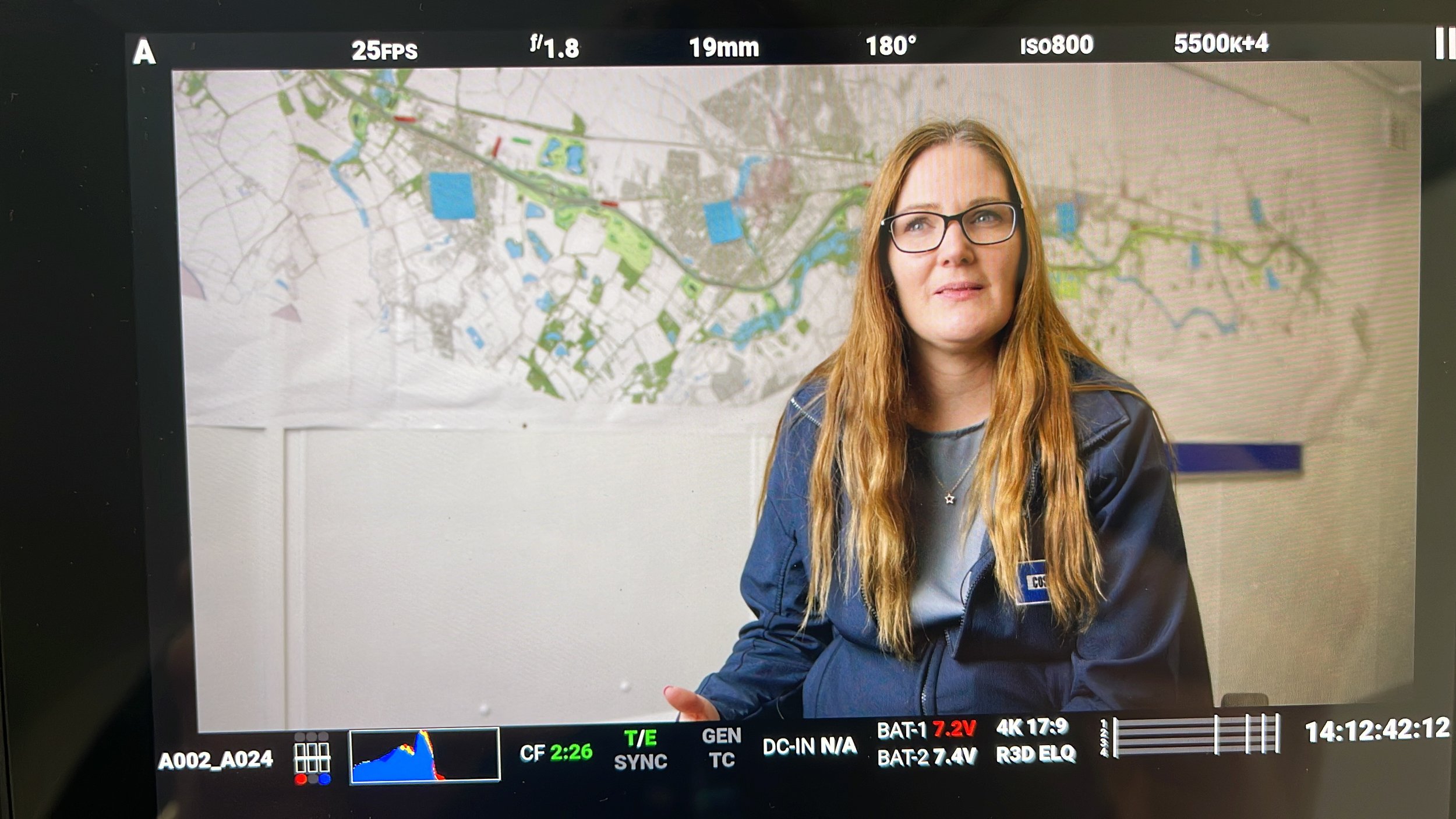 A woman with long red hair and glasses sitting in front of a map on a wall, with camera settings displayed at the top of the image.