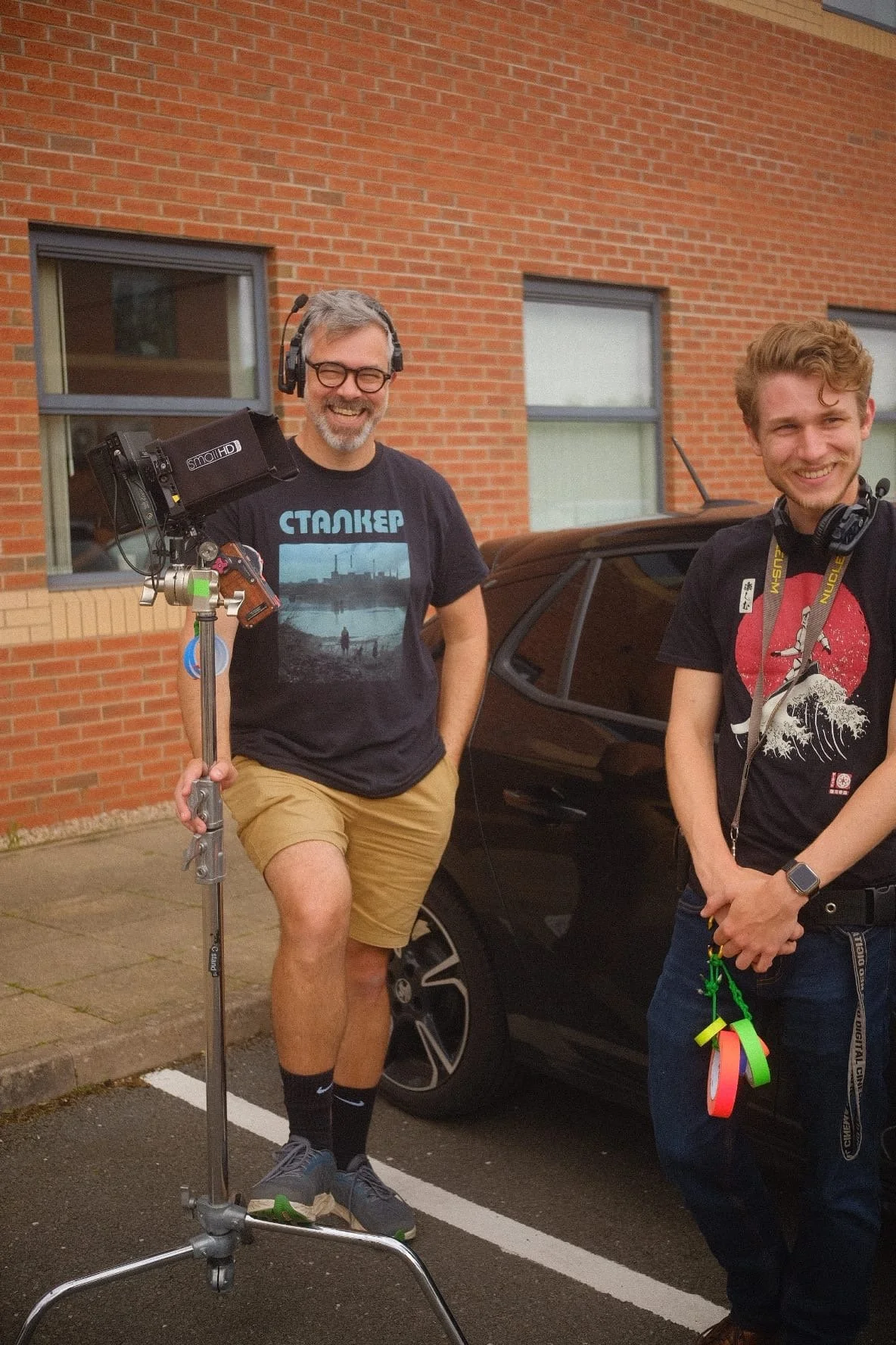 Two smiling men standing outdoors near a black car, with a brick building in the background. The man on the left holds a camera on a tripod, wearing a black t-shirt with a graphic, glasses, headphones, and shorts. The man on the right wears a black t