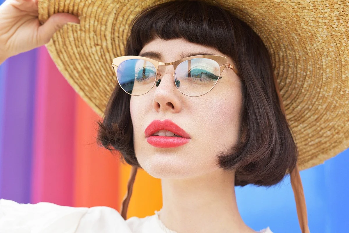 A woman wearing a large straw sunhat and sunglasses, with a colorful rainbow background.