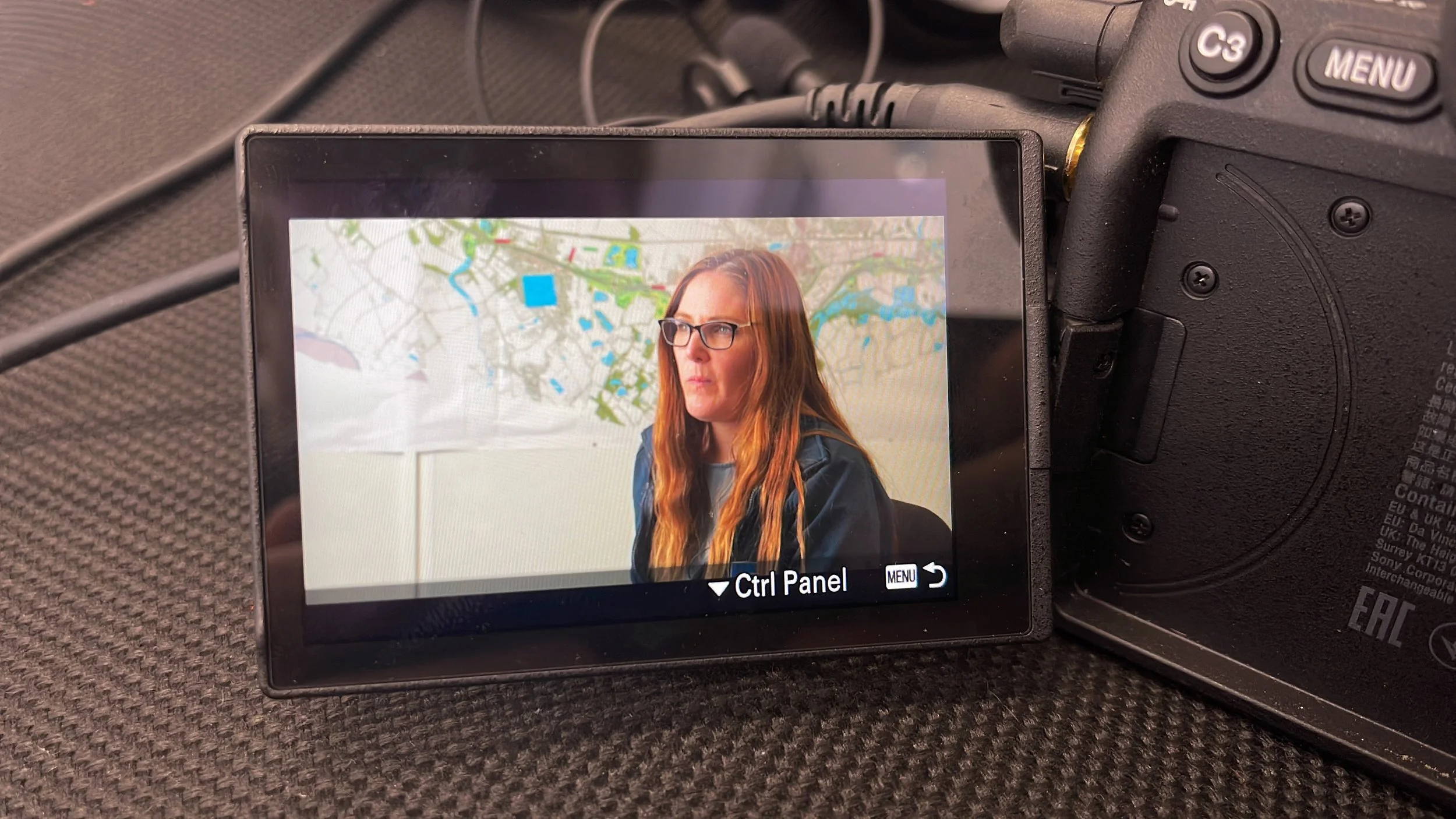 Camera screen displaying a woman with red hair and glasses in front of a map.