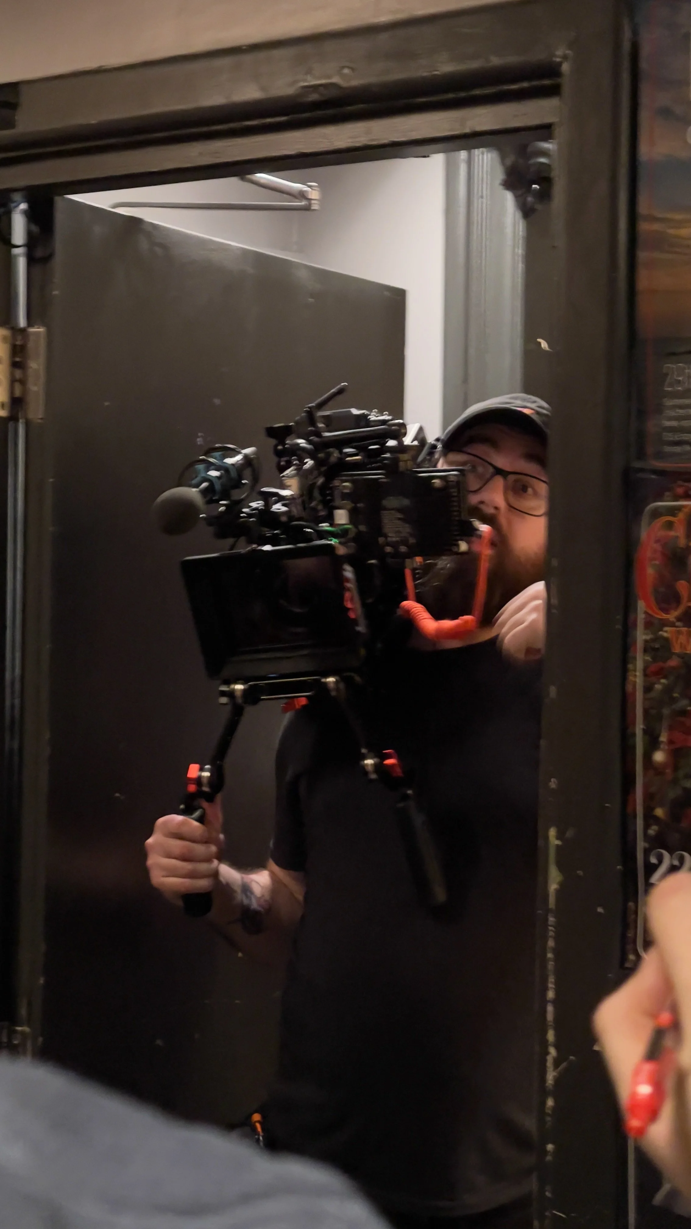 A man with glasses and a cap, holding a camera on a tripod, taking a selfie in a mirror, with a dark wall in the background.