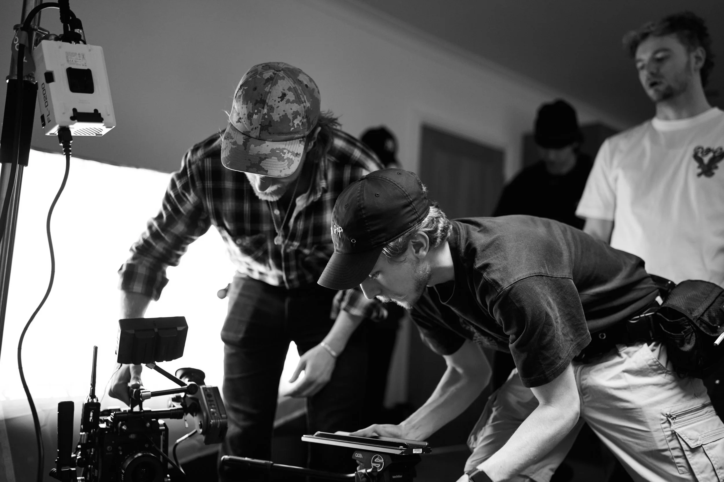 A group of people on a film or photography set working with professional equipment. One person is crouched down, operating a camera, while others are looking on and assisting. The scene appears to be set indoors.