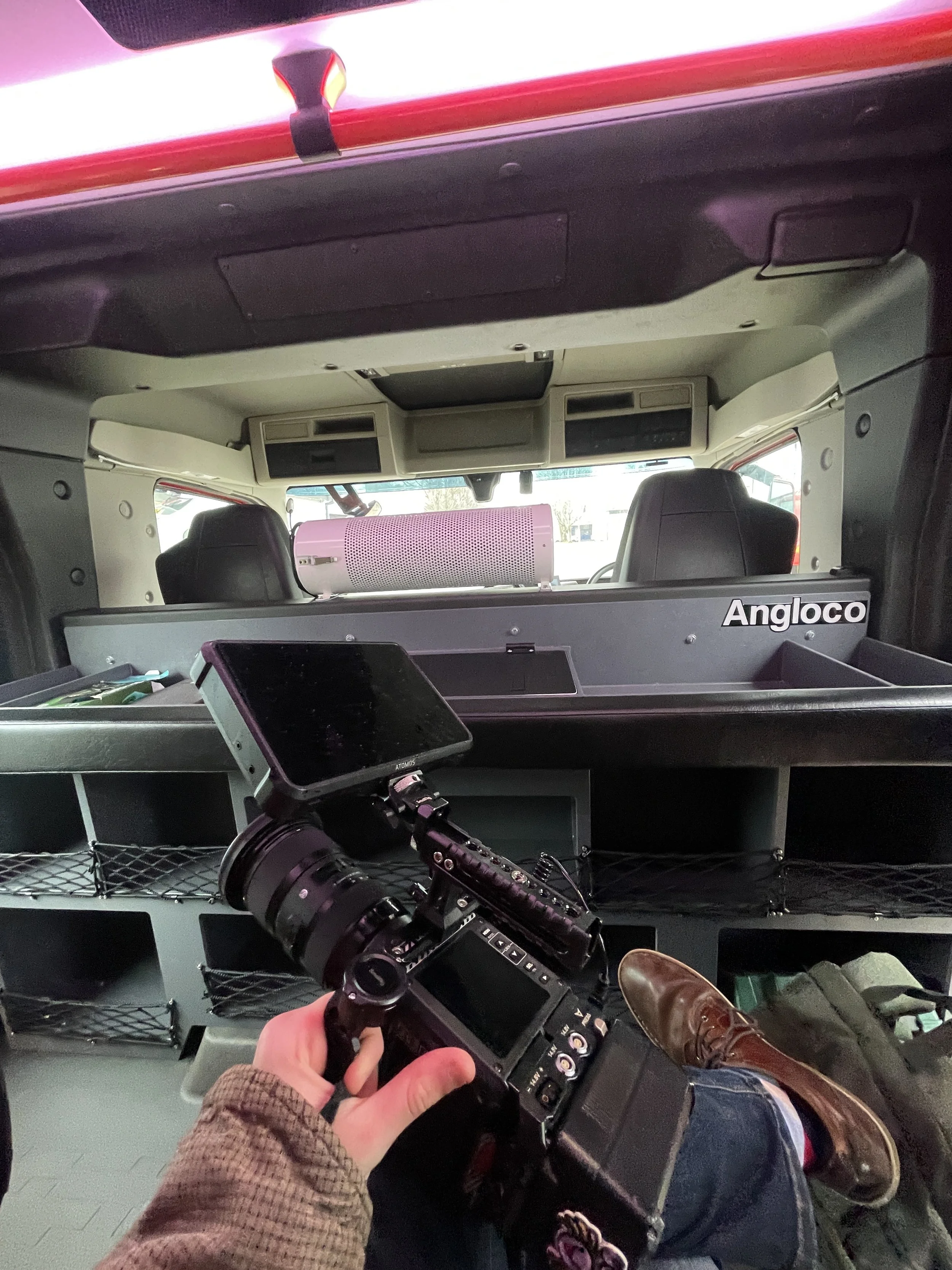 Inside the back of a police vehicle, featuring equipment and a person's legs with boots, a hand holding a remote control, and various electronic devices and storage compartments.