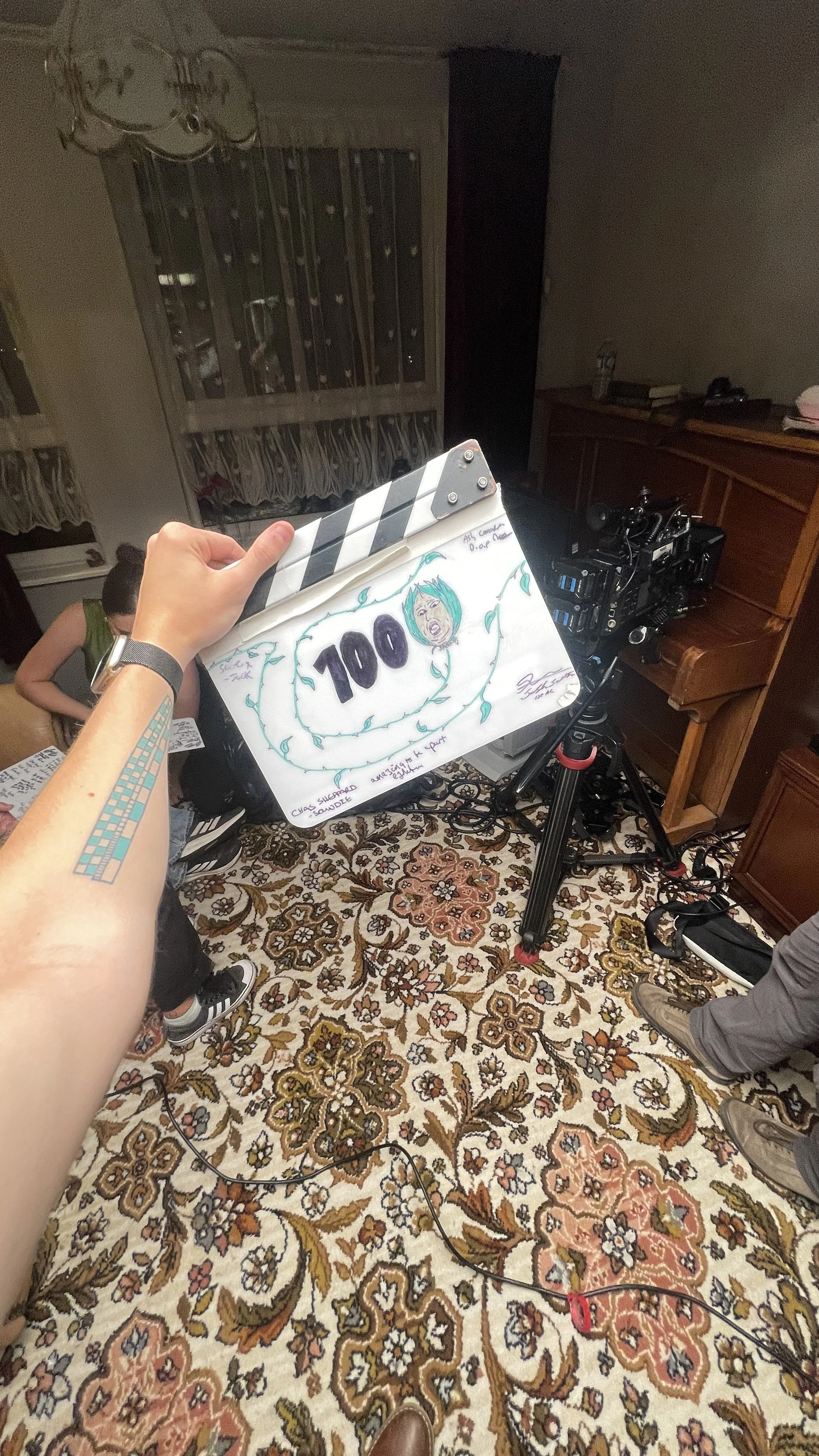 A person holding a clapperboard with a drawing of a face and the number 100 written on it in a room with a patterned rug, a camera on a tripod, and other people and furniture visible in the background.