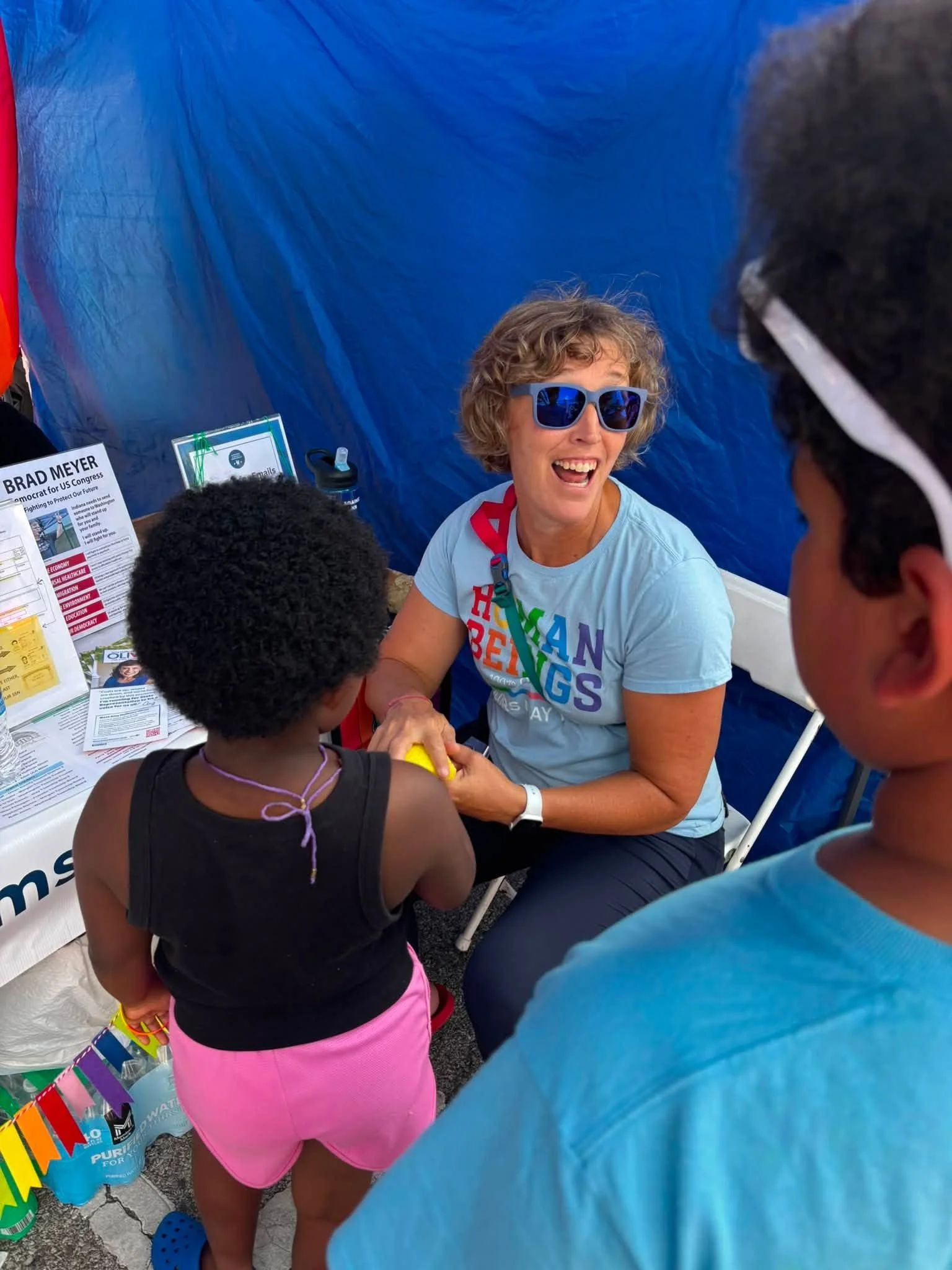 A woman with curly hair and sunglasses smiling while shaking hands with two children at an outdoor booth, which has informational signs and rainbow-colored decorations.