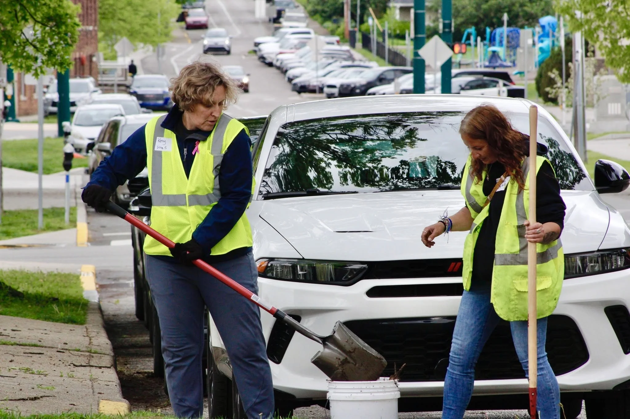 Two women in yellow safety vests working on a white car in a parking lot, with trees and parked cars in the background.