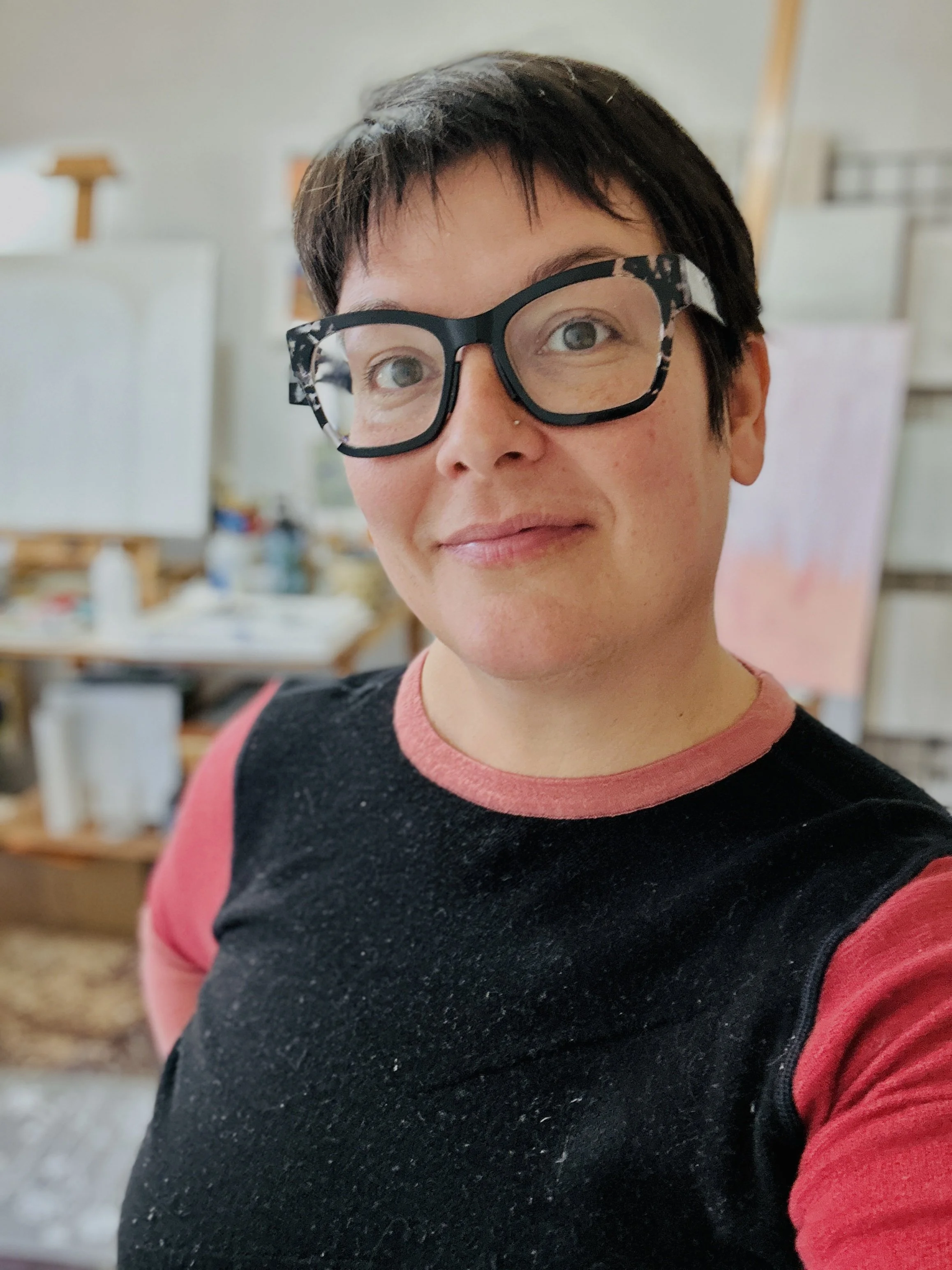 A woman with short dark hair, glasses with a black and white pattern, and a nose piercing, taking a selfie in an art studio. She is wearing a black and red shirt, and the background includes art supplies, easels, and artwork.