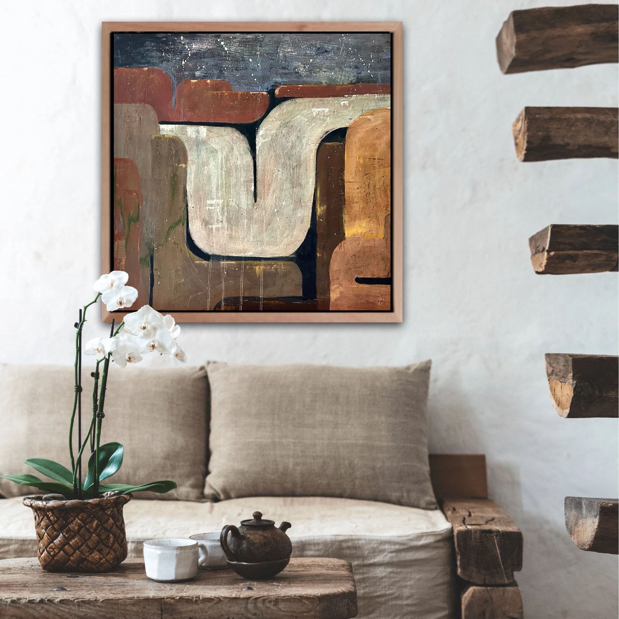 A living room with a beige couch, beige cushions, a rustic wooden coffee table with a white orchid in a wicker pot, a teapot, and a small cup. A framed abstract painting with earth tones hangs on the white wall.