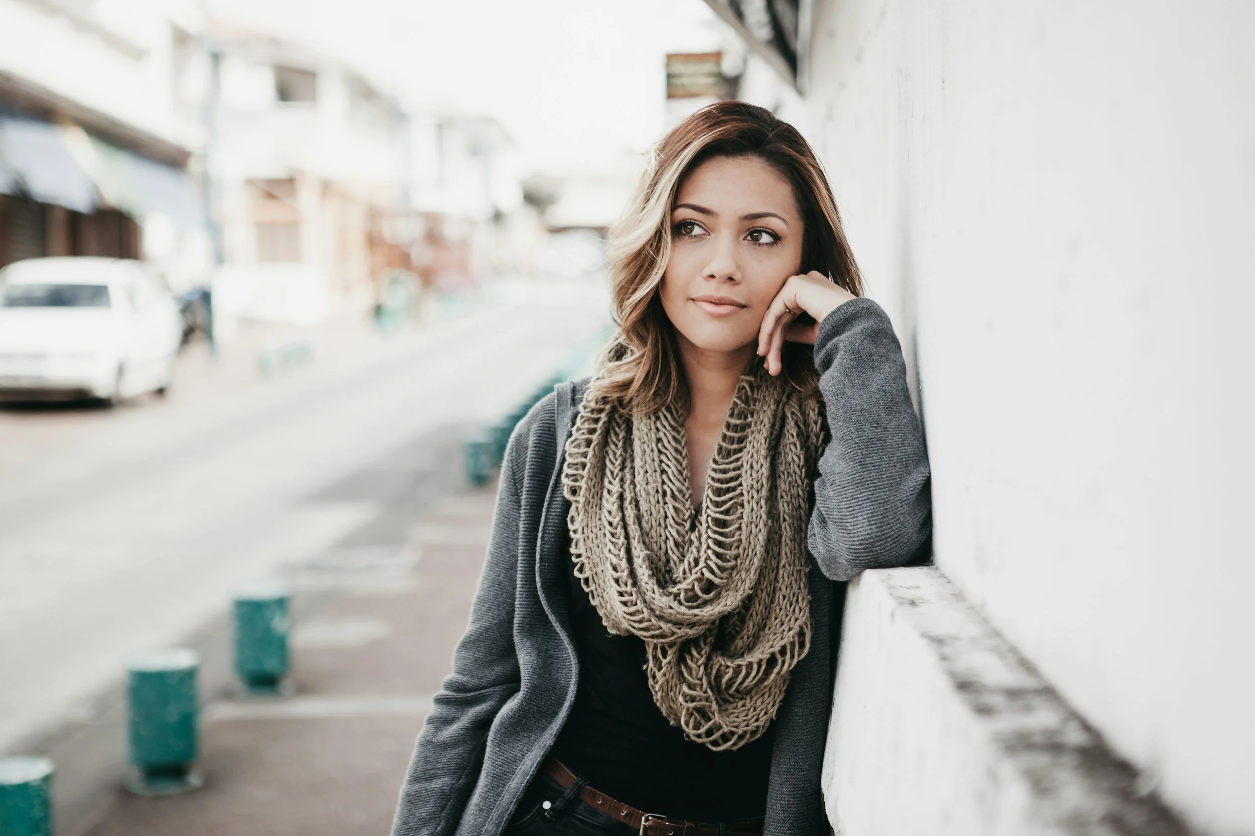 A woman with brown hair and light skin leaning against a white wall on a city street, wearing a gray coat and beige knit scarf.