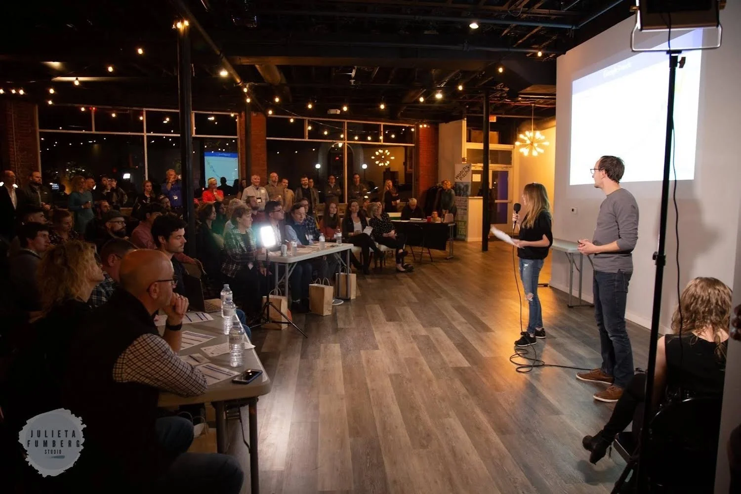 A presentation or speech taking place in a well-lit, modern indoor venue with a large audience. Two speakers stand at the front near a big screen, one holding a microphone. The audience is seated at tables and some are standing in the back, attentively watching the speakers.