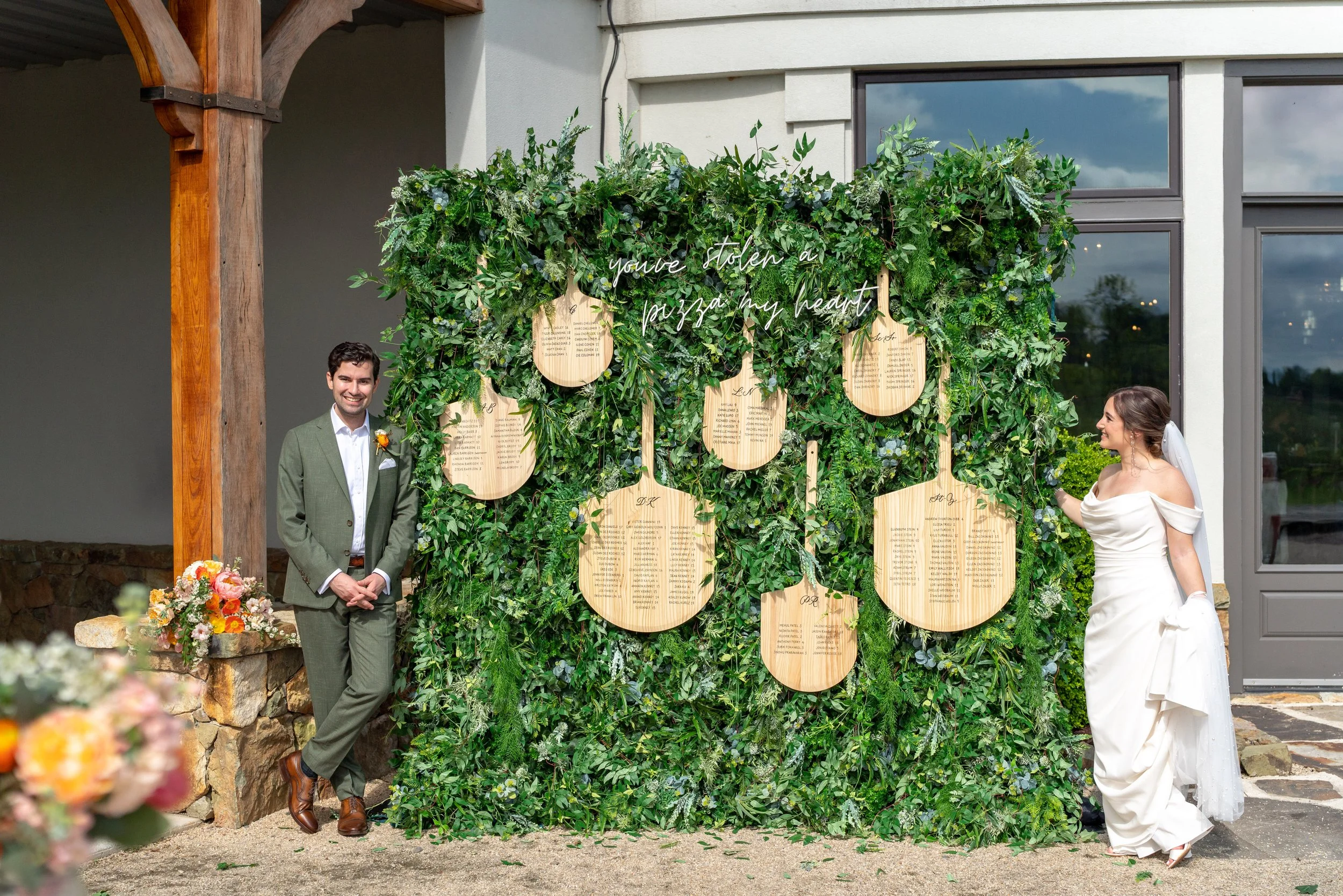 Creative wedding seating chart made from wooden pizza paddles with custom calligraphy, displayed on a greenery wall with neon sign that reads “you’ve stolen a pizza my heart”