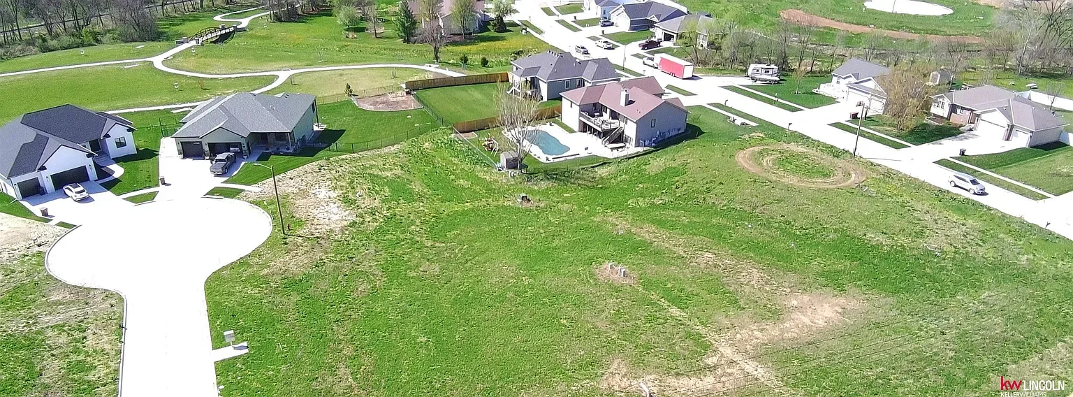 Aerial view of a residential neighborhood with single-family homes, a green park area, and a few empty lots. Some houses have driveways and garages, with one house featuring a swimming pool in the backyard.