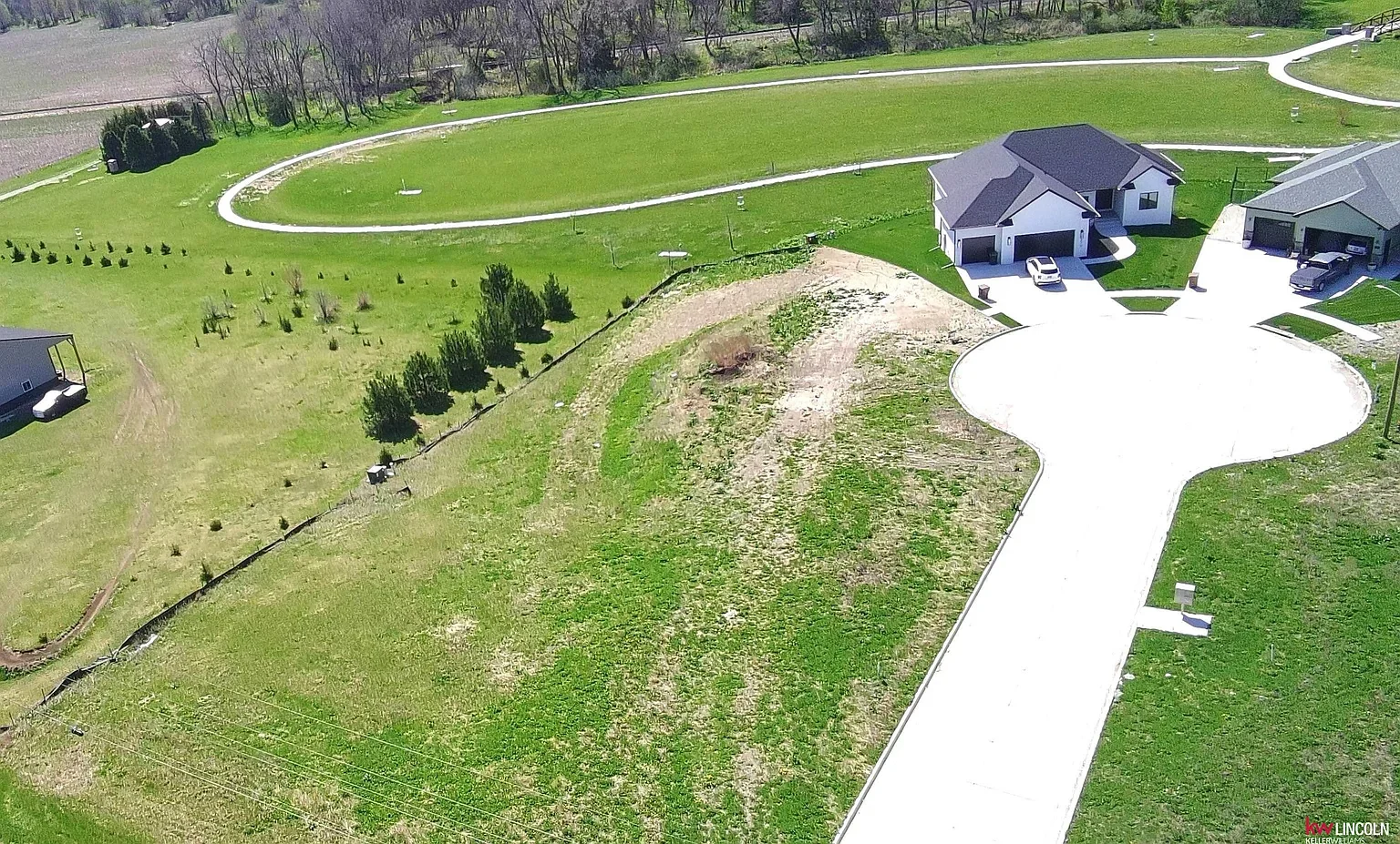 Aerial view of a suburban neighborhood showing houses, driveways, and green lawns, with a dirt hill and a winding white pathway or irrigation system in the background.