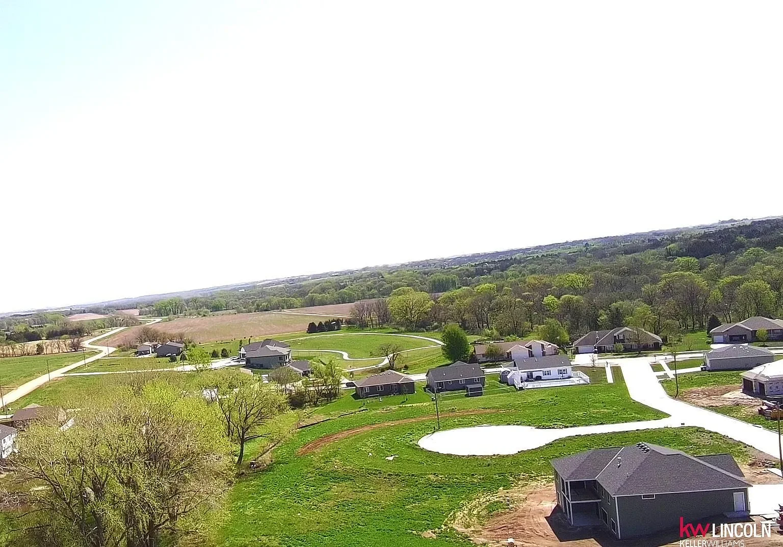 A scenic aerial view of a suburban neighborhood with multiple houses, green lawns, some trees, and winding roads, under a clear sky.