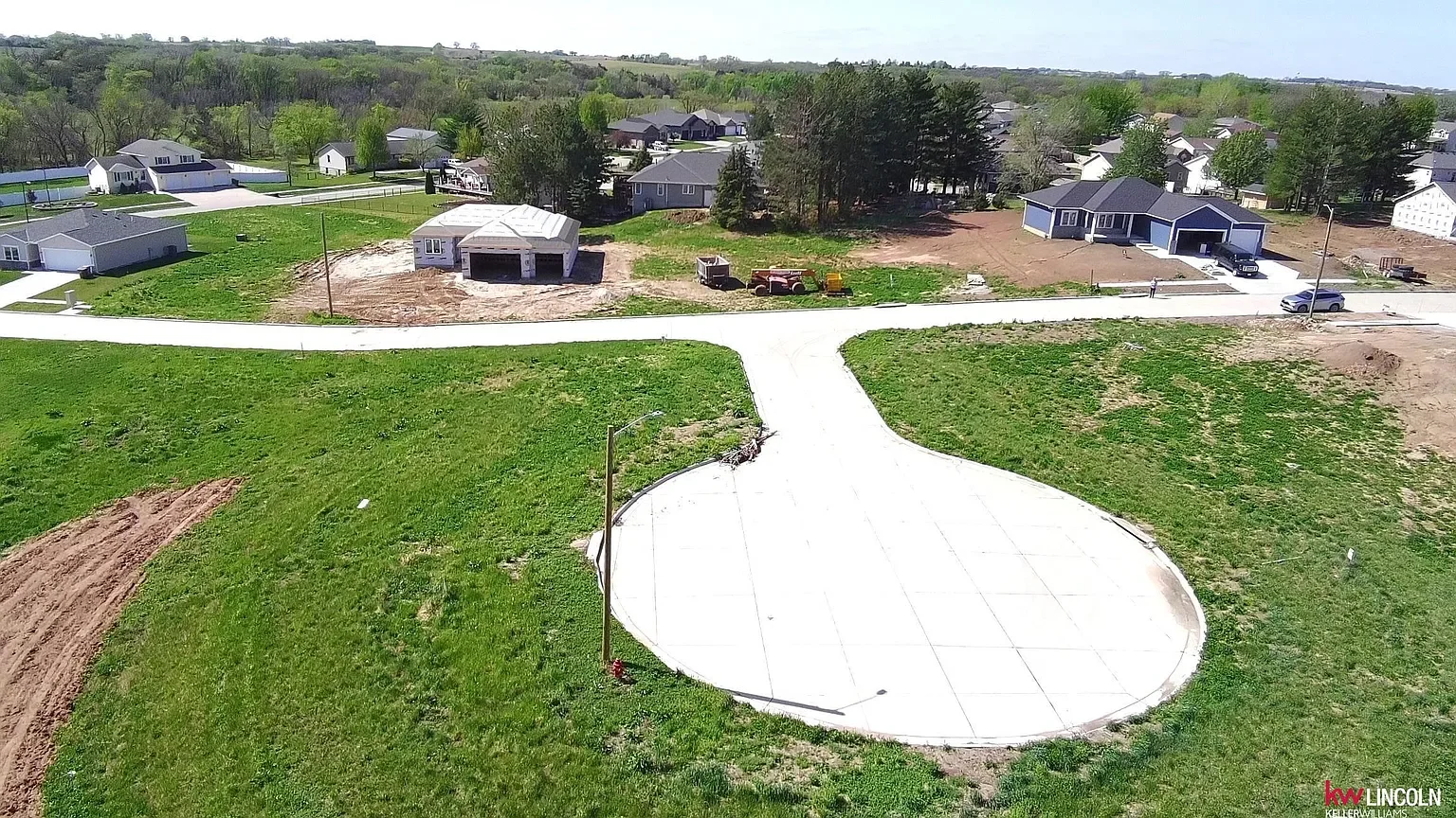 A suburban residential area with some houses under construction, a curved concrete driveway or pad, and patches of green grass.