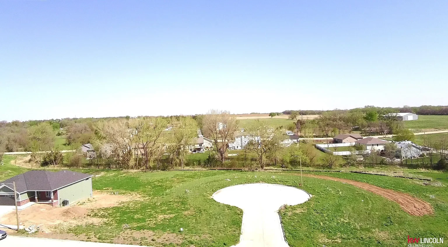 A rural landscape with a few houses, trees, and green fields under a clear blue sky.
