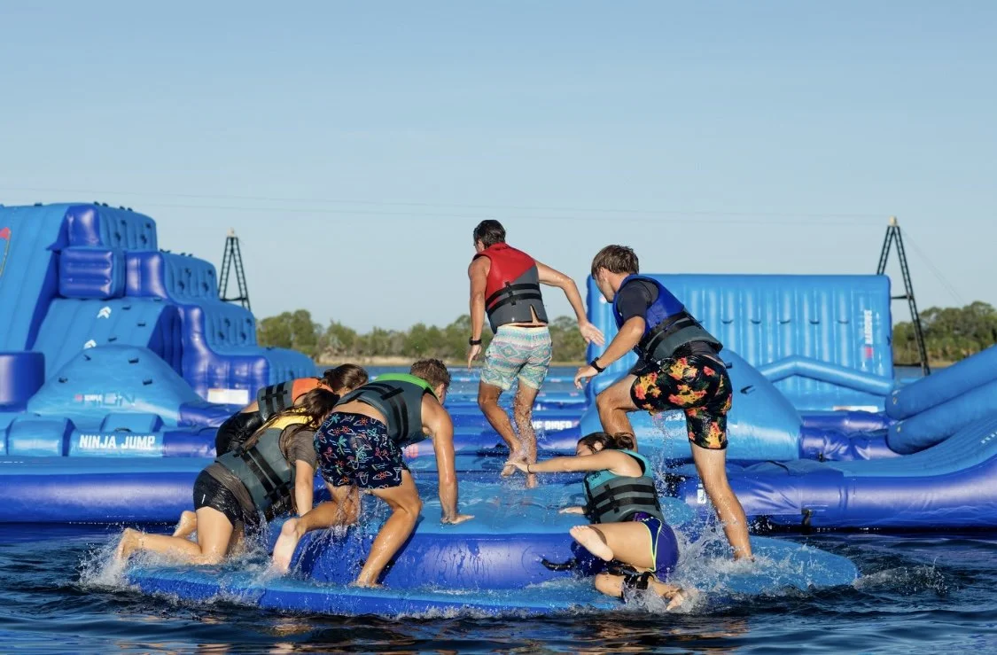 People enjoying a jump on an inflatable water obstacle course in a lake or water park, wearing life jackets.