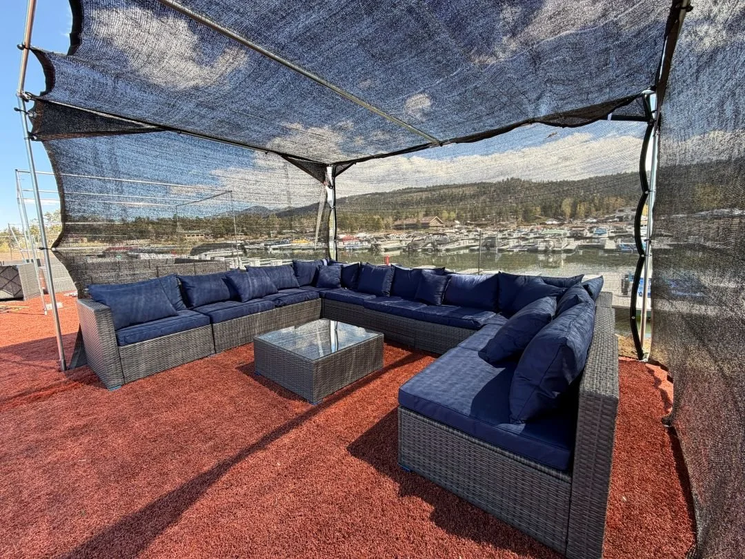 Outdoor seating area with a large L-shaped wicker sectional sofa with blue cushions, a glass-topped wicker coffee table, under a mesh canopy with a view of a marina and hills in the background.
