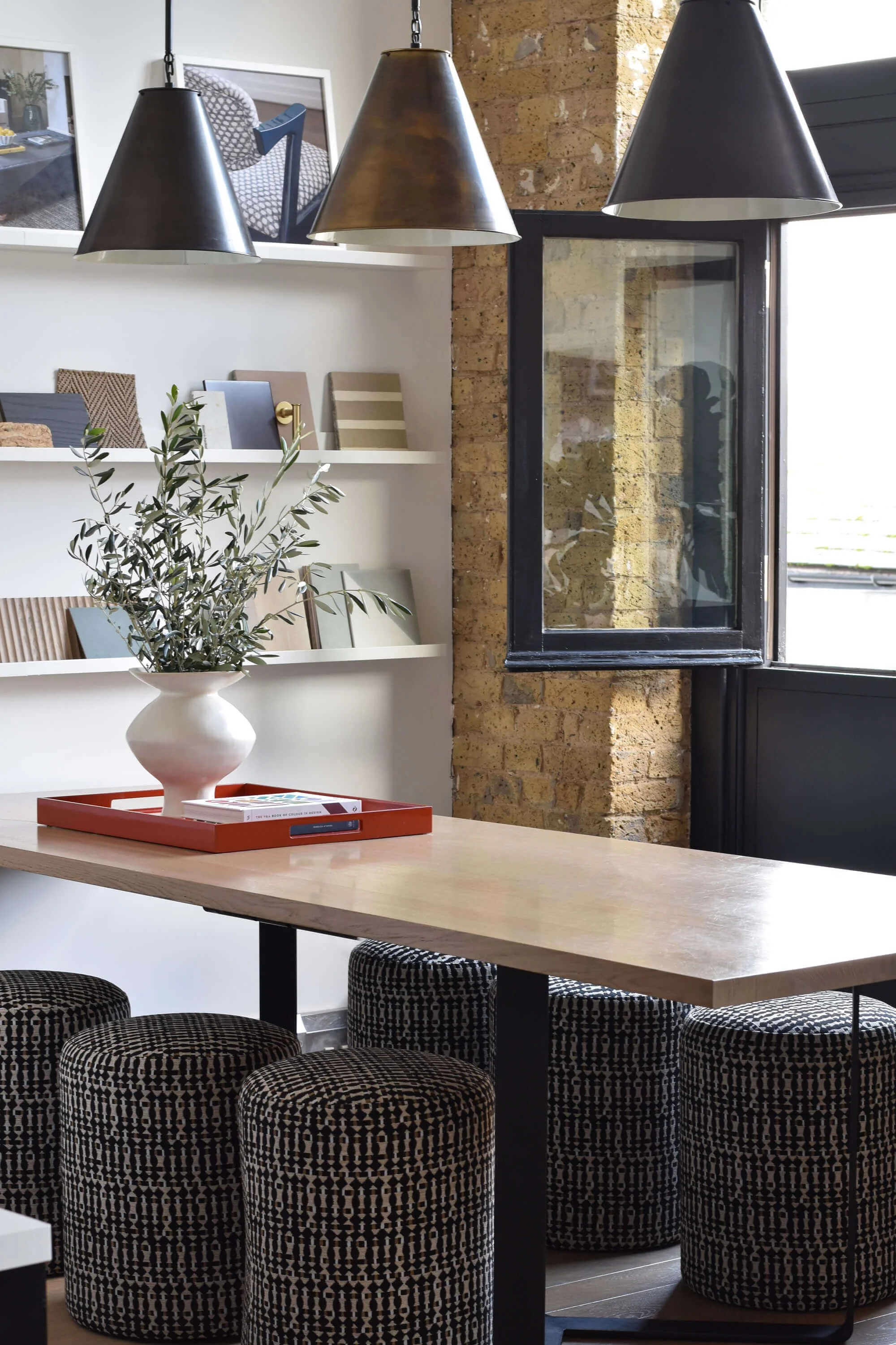 Interior of Th2 Designs design studio with a wooden table, black and white patterned upholstered stools, a white vase with greenery on a tray, pendant lights, a wall with picture frames, and a large window with a brick wall outside.
