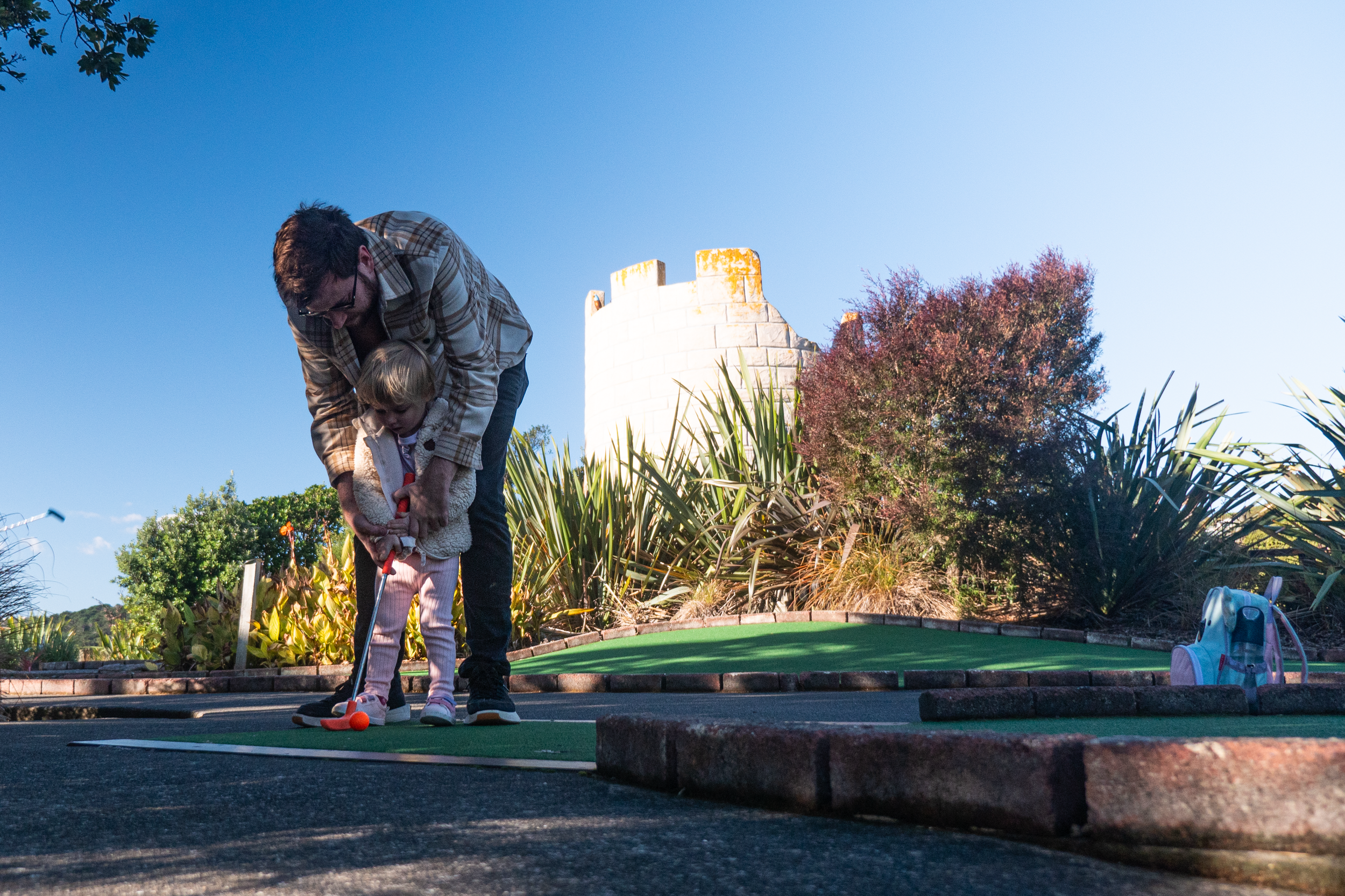 A man wearing a checked jacket and sunglasses is helping his young daughter play golf at the adventure golf course. The sky is blue and in the background is a white brick castle tower.