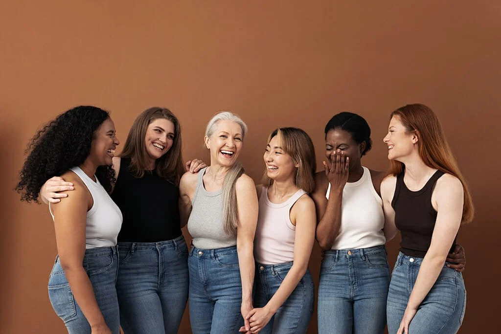 Group of six women laughing and enjoying each other's company against a plain brown wall.