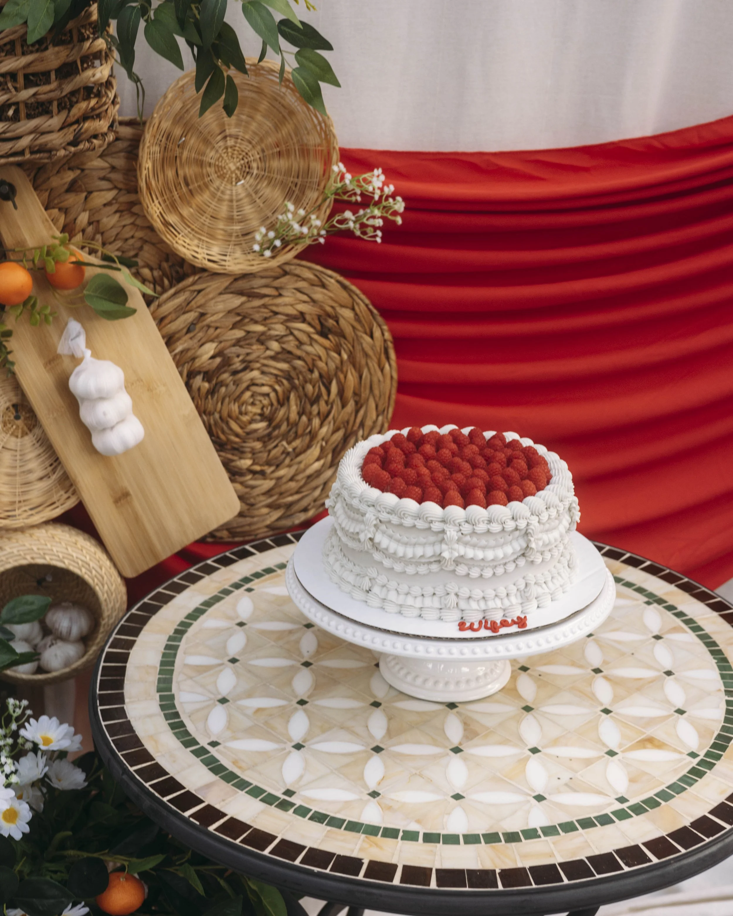 A white cake decorated with red berries on top, on a white cake stand, placed on a round tiled table with a cream and green pattern. The background has woven baskets, green foliage, and red fabric draping.