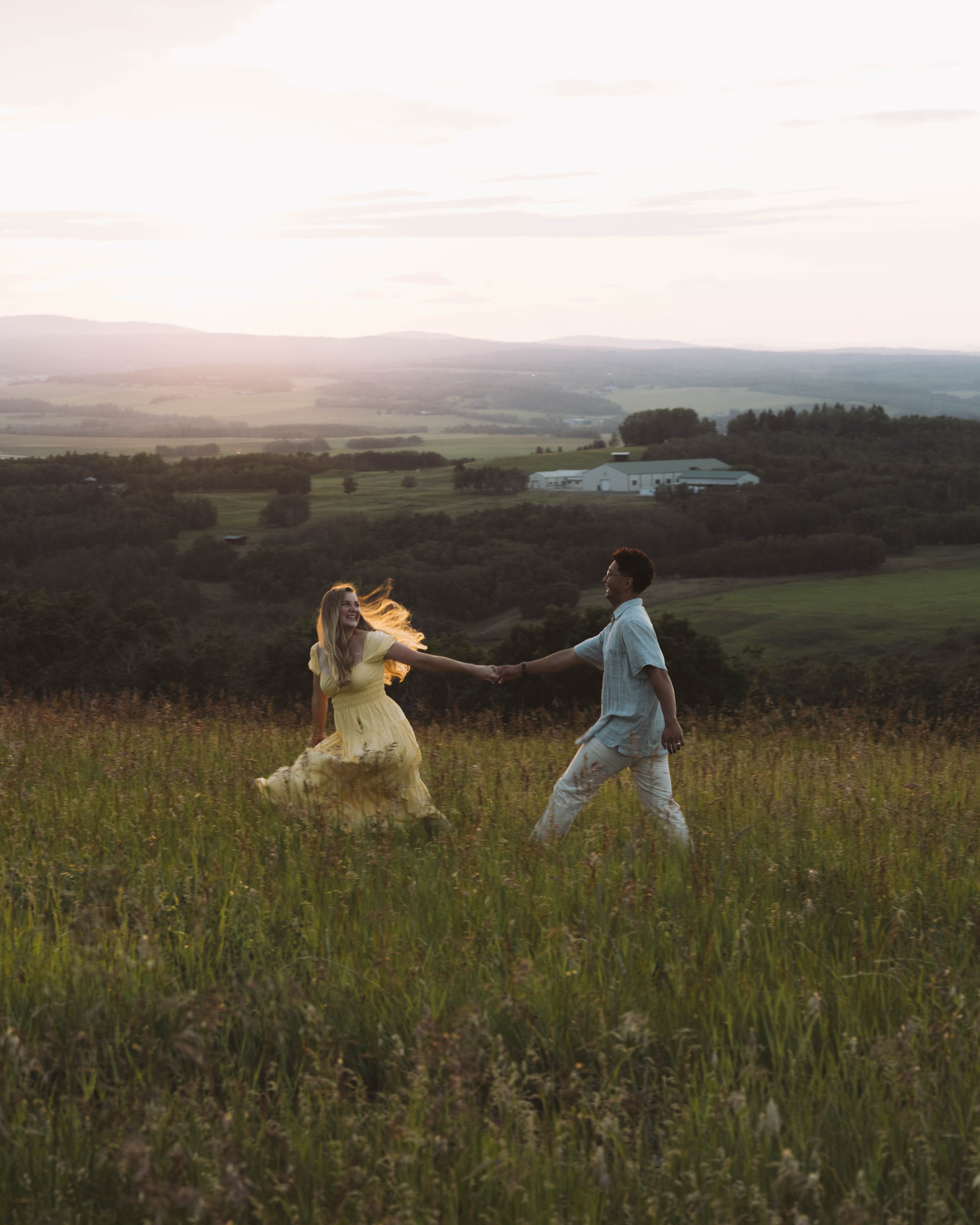 A couple holding hands and running through a grassy field at sunset, with a rural landscape and farm buildings in the background.