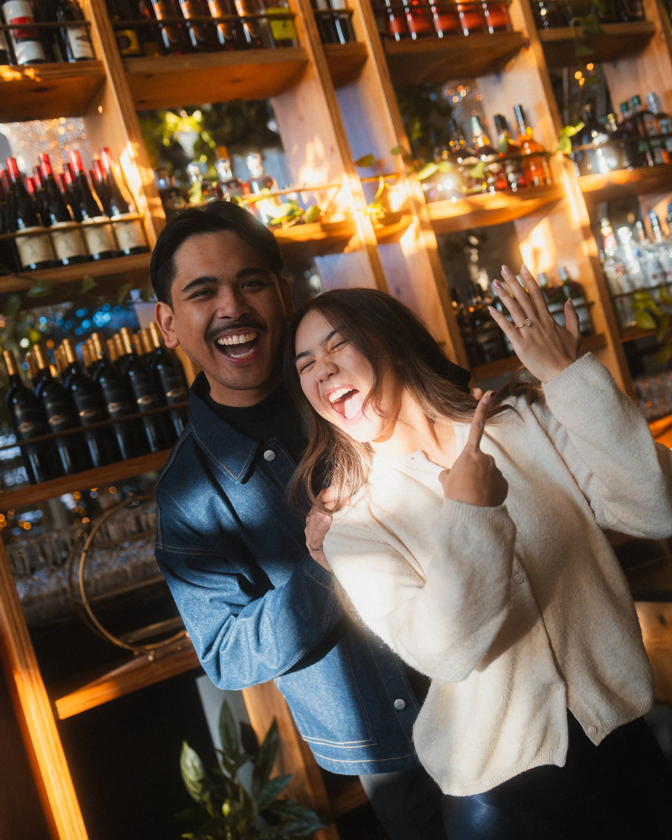 Happy couple celebrating in a bar with wine bottles on wooden shelves behind them.