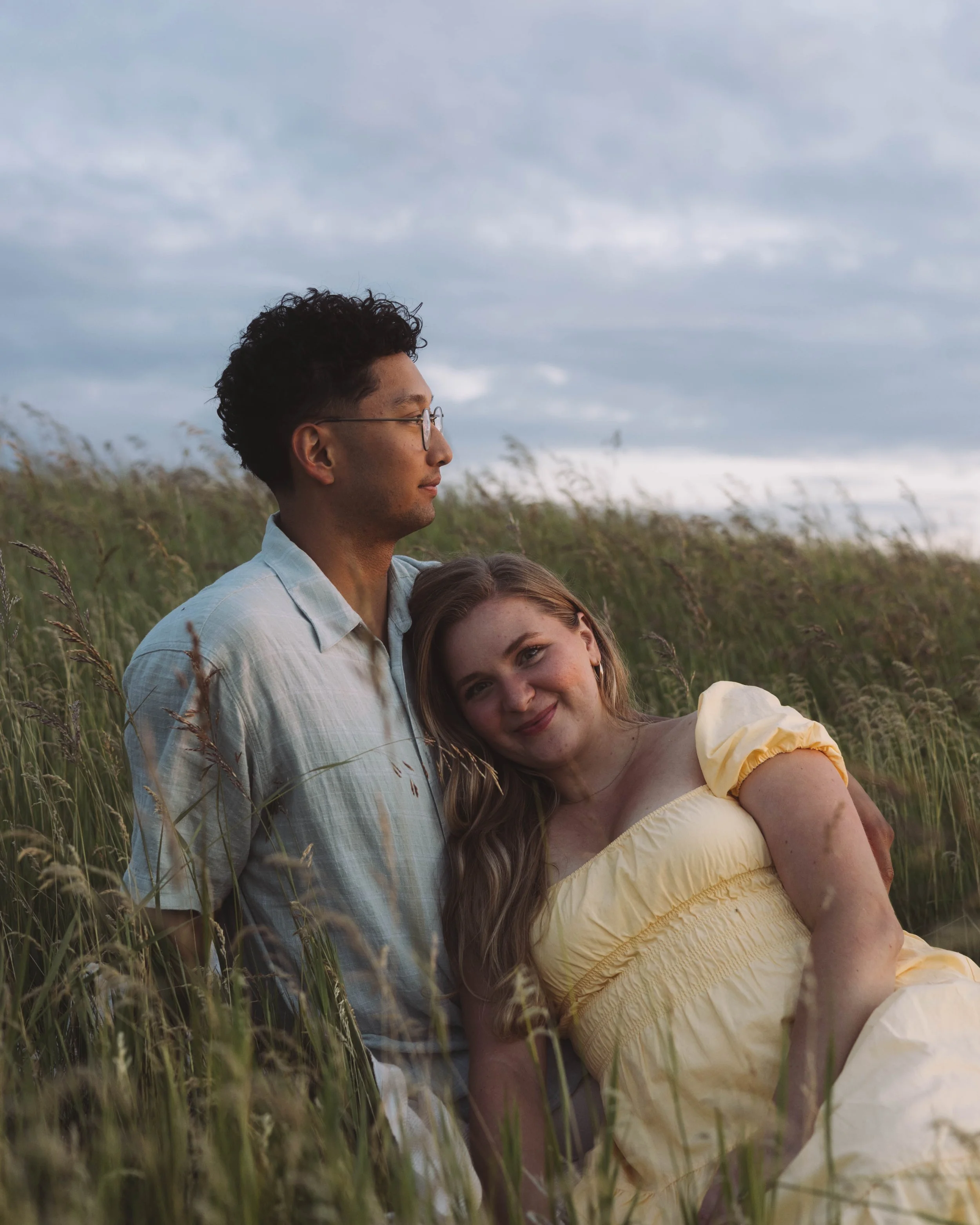 A young couple sitting in tall grass outdoors at sunset, with the woman resting her head on the man's shoulder and smiling, while the man looks into the distance.