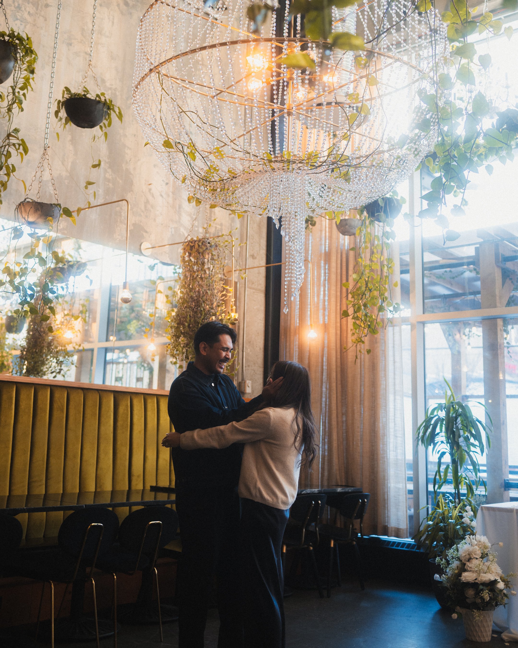 A couple dancing in a warmly lit, decorated indoor space with a large chandelier, greenery, and warm curtains.